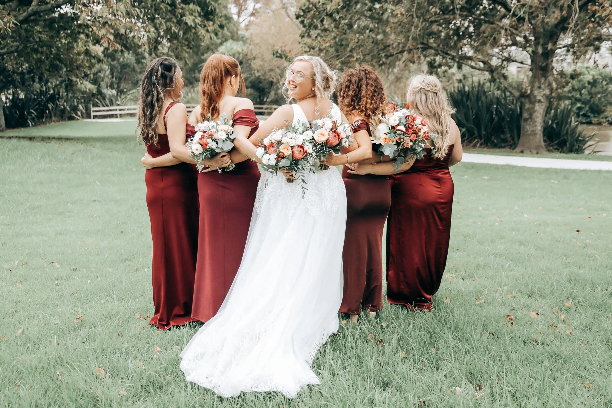 A bride in a white wedding gown walking in a grassy park, surrounded by five bridesmaids in matching dark red dresses, holding bouquets of flowers, with trees in the background.