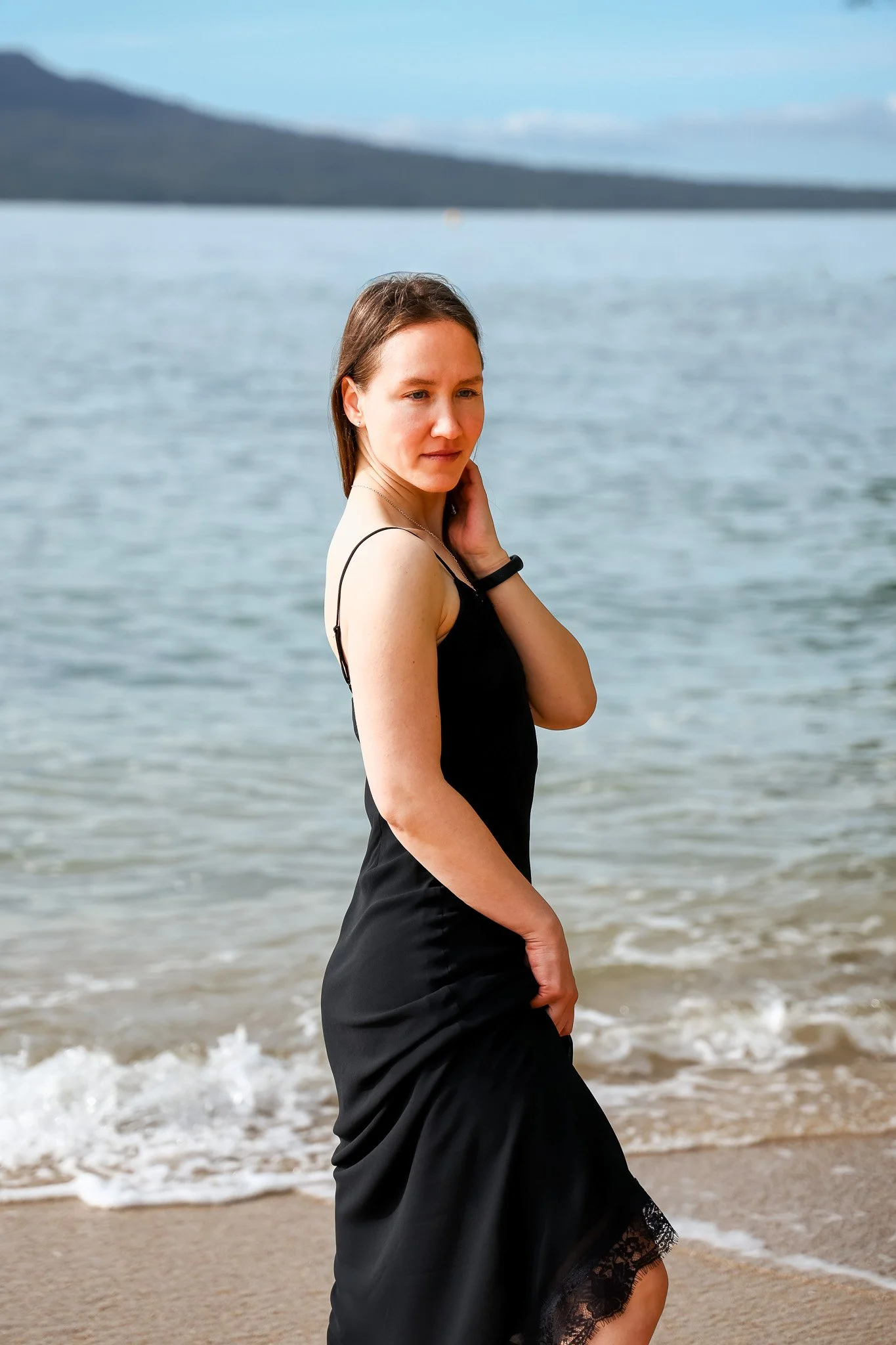 A woman in a black dress standing at the beach with water and distant mountains in the background.