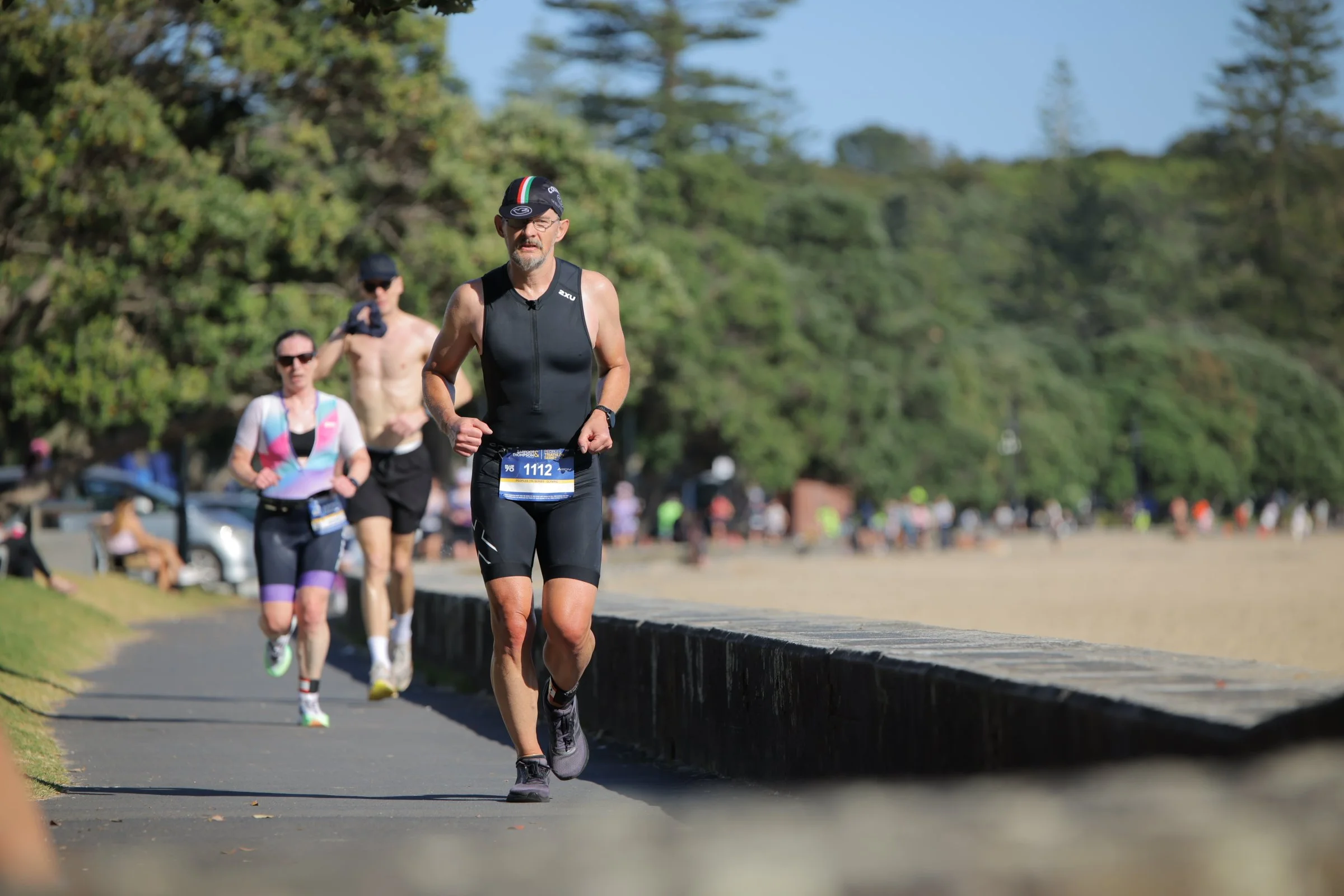 A man running in a race or marathon, wearing a black sleeveless athletic top, black shorts, a cap with an Italian flag stripe, and sunglasses, with a bib number 1112. He is on a paved path in a park, with trees and other runners visible in the backgr