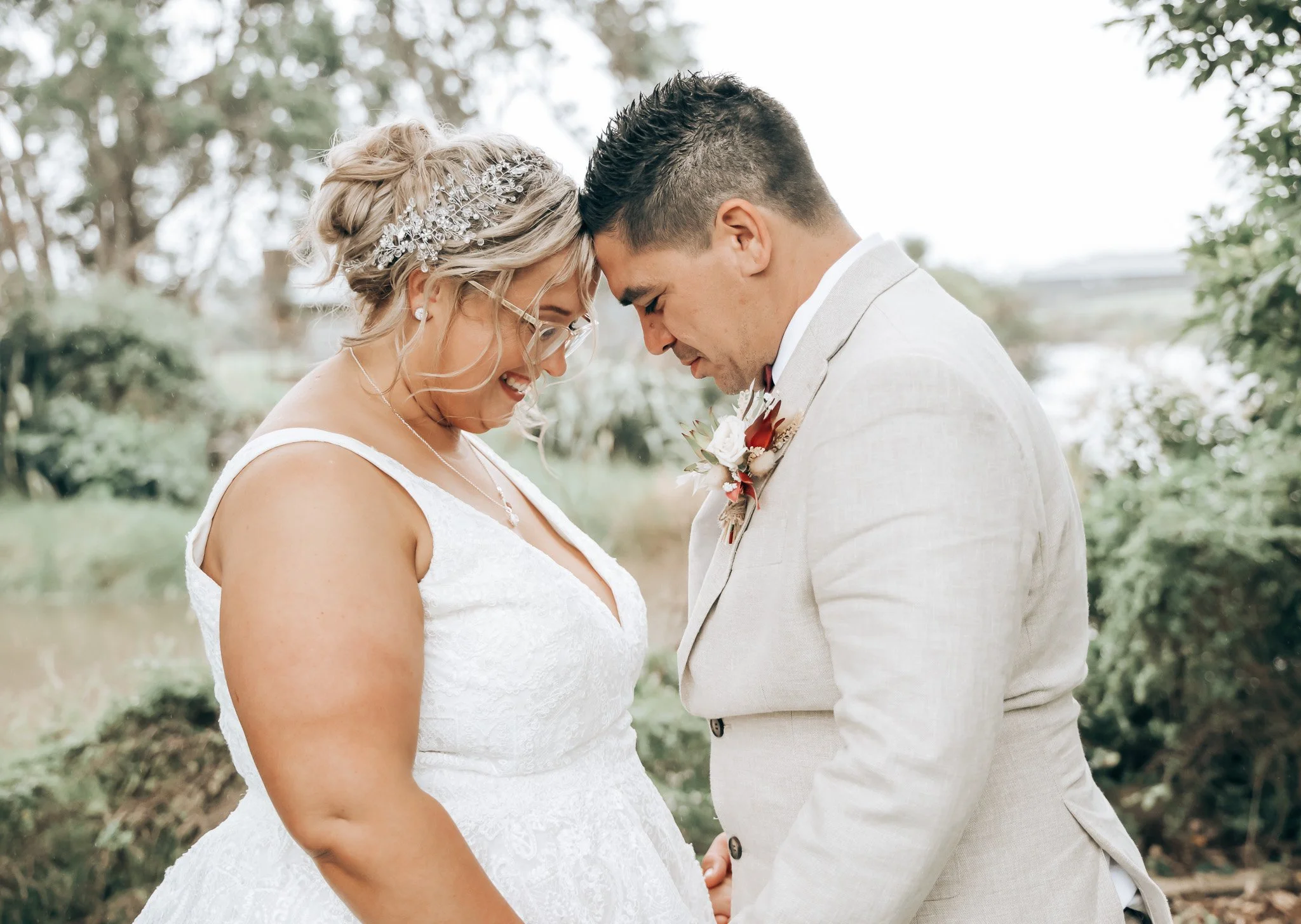 A couple dressed in wedding attire sharing a tender moment outdoors, holding hands and touching foreheads with eyes closed, surrounded by trees and natural scenery.