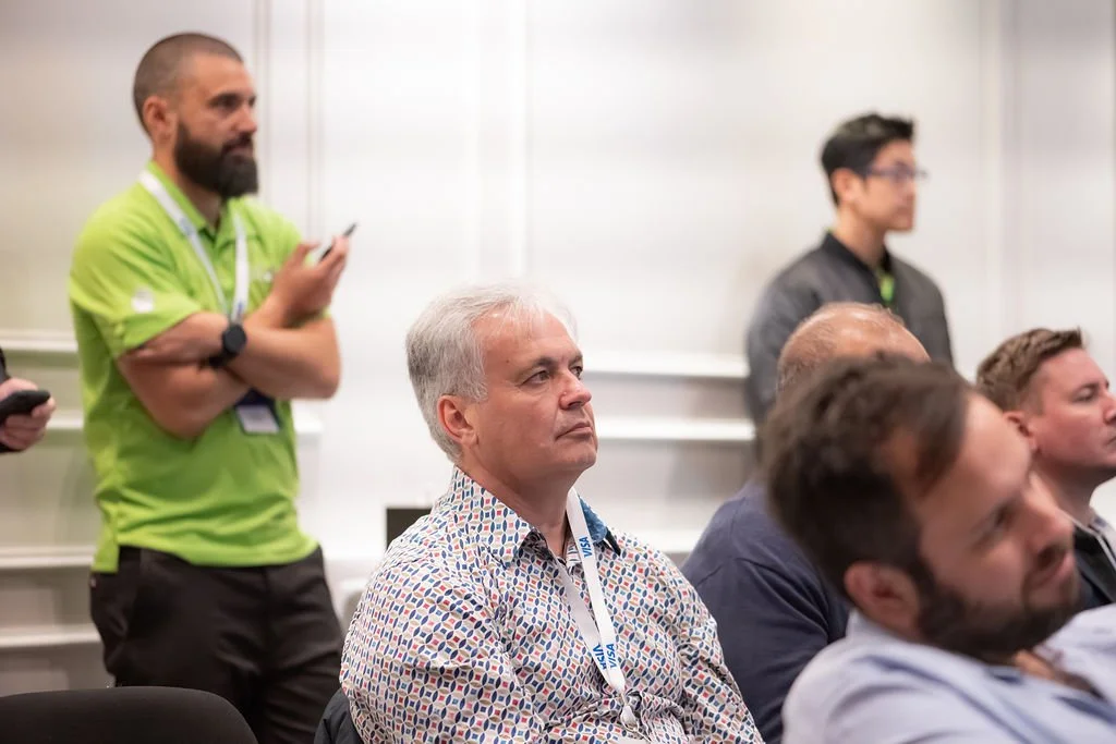 Group of men attending a conference, sitting and listening to a speaker. Two men are standing, one wearing a green shirt and another in a dark jacket, both holding a phone.