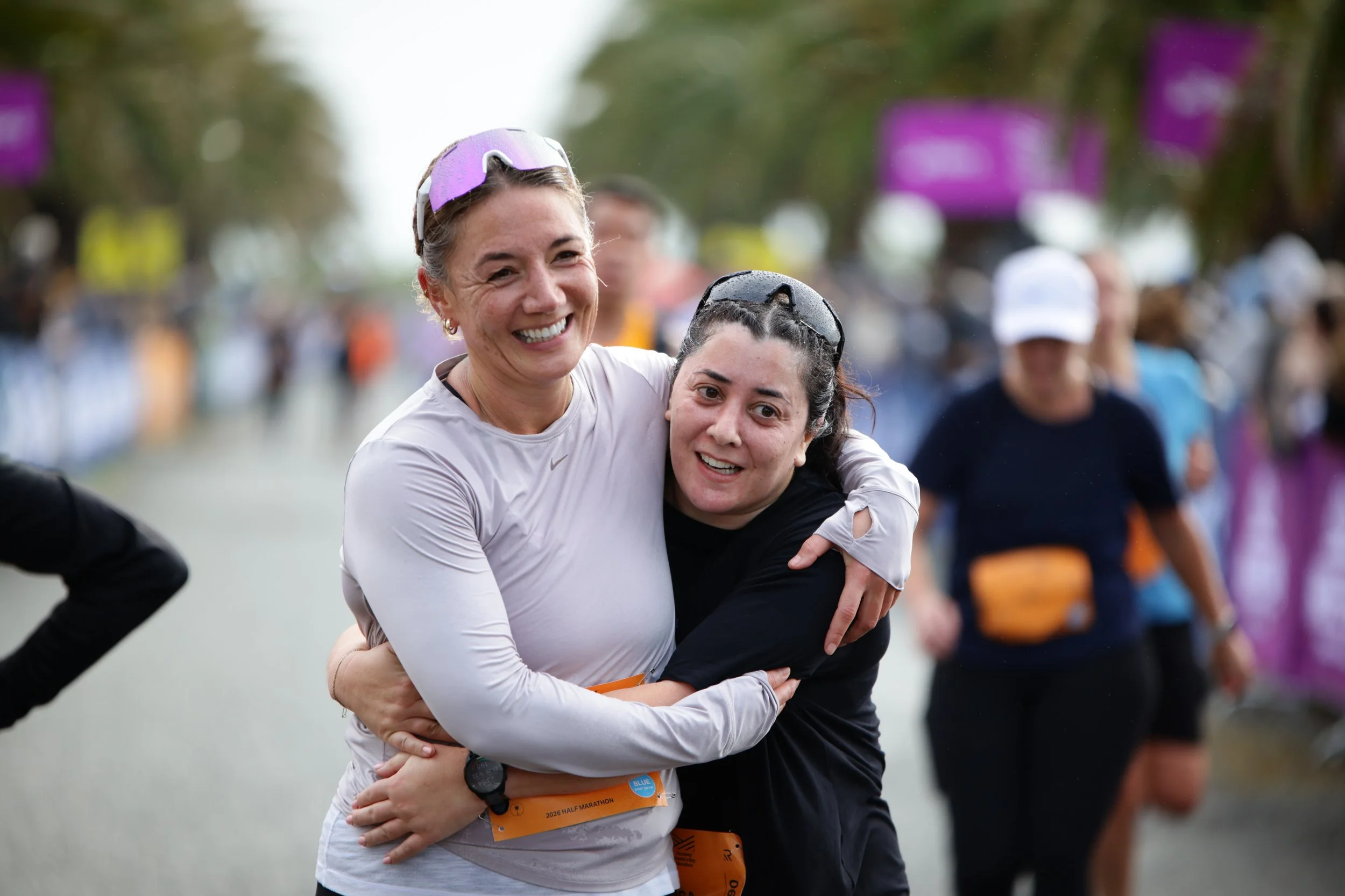 Two women hugging and smiling at a marathon event, with other participants and purple signs visible in the background.