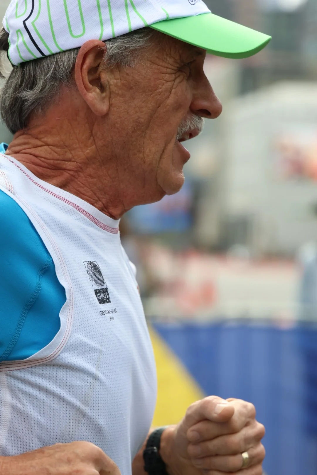 A close-up side profile of an older man with gray hair and a mustache, wearing a white and blue athletic shirt, a white hat with green accents, and a black wristwatch, appears to be running or exercising.