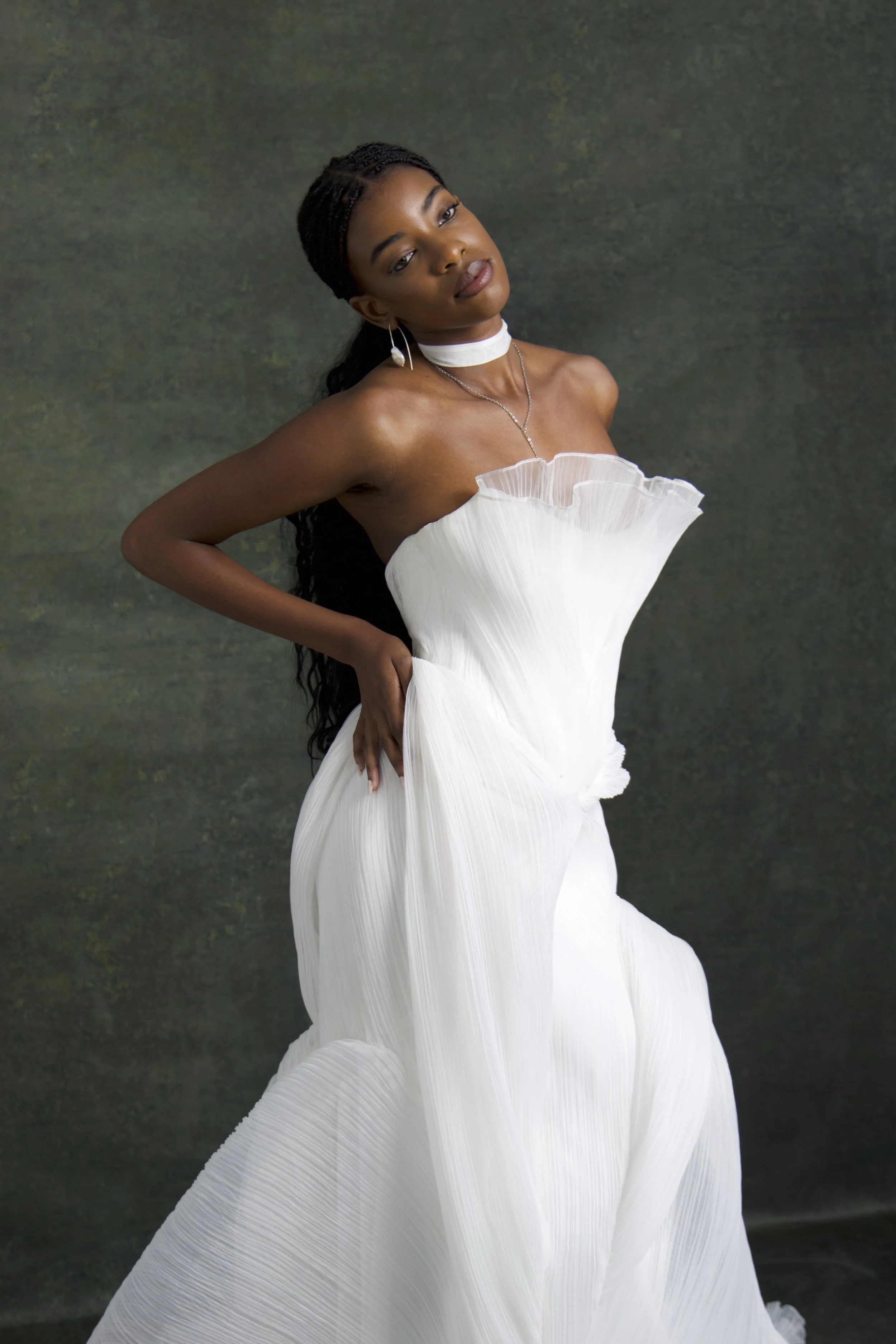 A woman with dark skin and long braided hair wearing a white off-shoulder dress with ruffled fabric, a white choker, and earrings, standing against a dark green textured background.