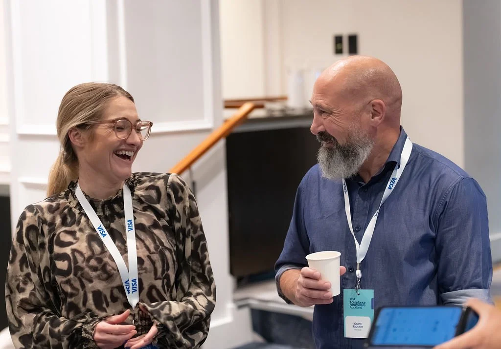 Two people, a woman with glasses and a man with a beard and a lanyard, are smiling and talking at an indoor event, with the woman wearing a leopard print top and the man holding a paper cup.