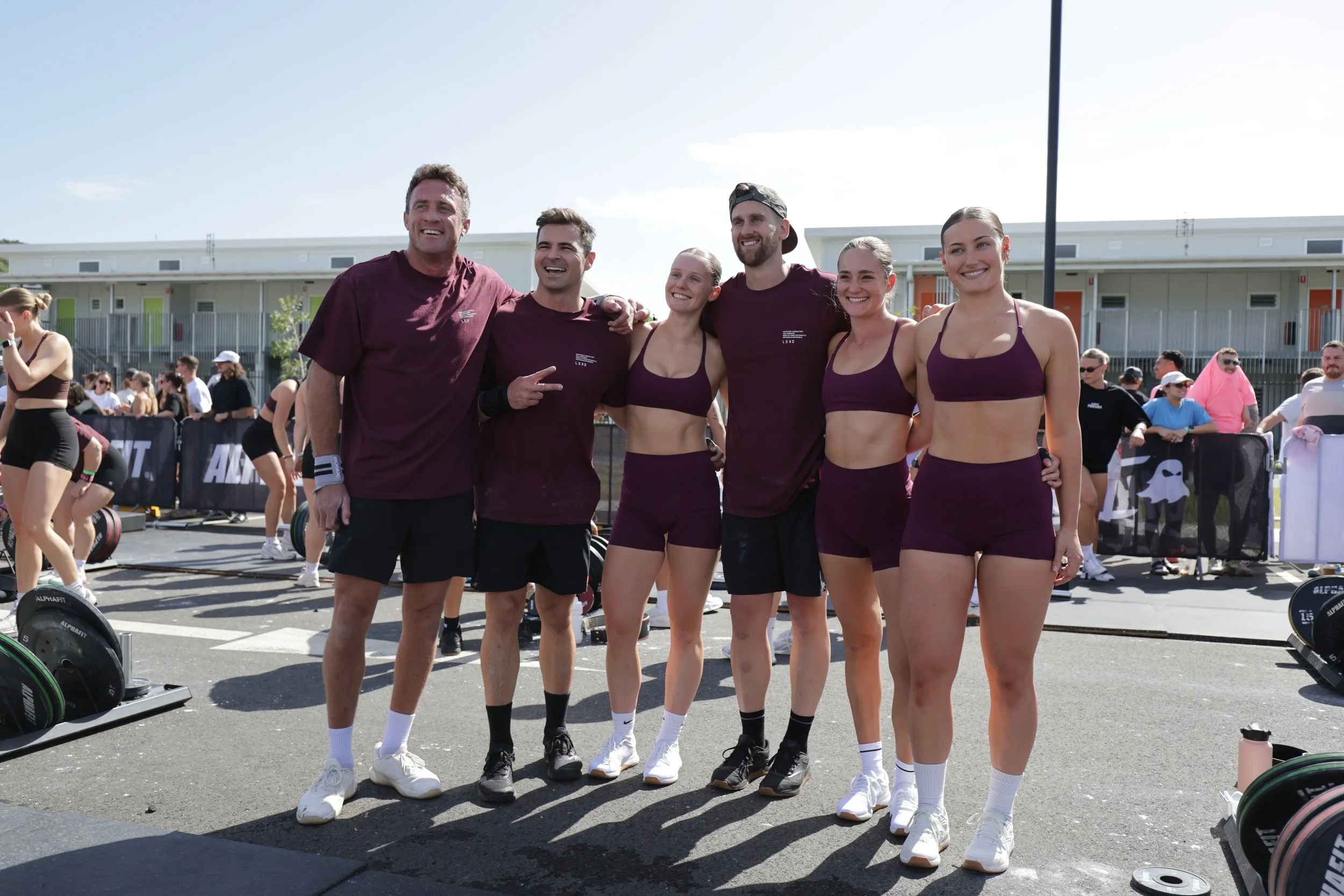 Group of six young athletes, three men and three women, standing outside at a sports event, smiling and posing for a photo, with weightlifting equipment and other spectators in the background on a sunny day.