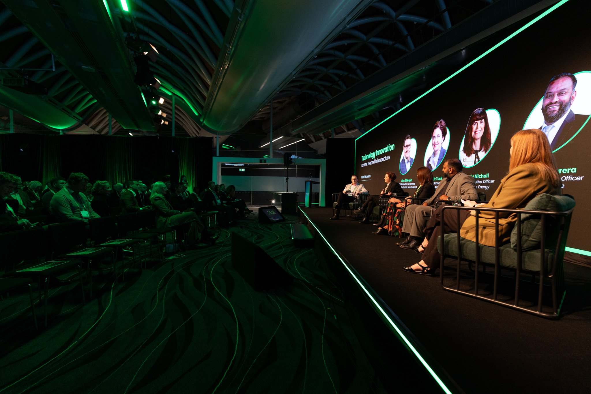 A large conference room with a panel of speakers on stage and an audience listening. The stage has a big screen displaying speakers' names and titles, illuminated with green accent lighting on the ceiling and walls.