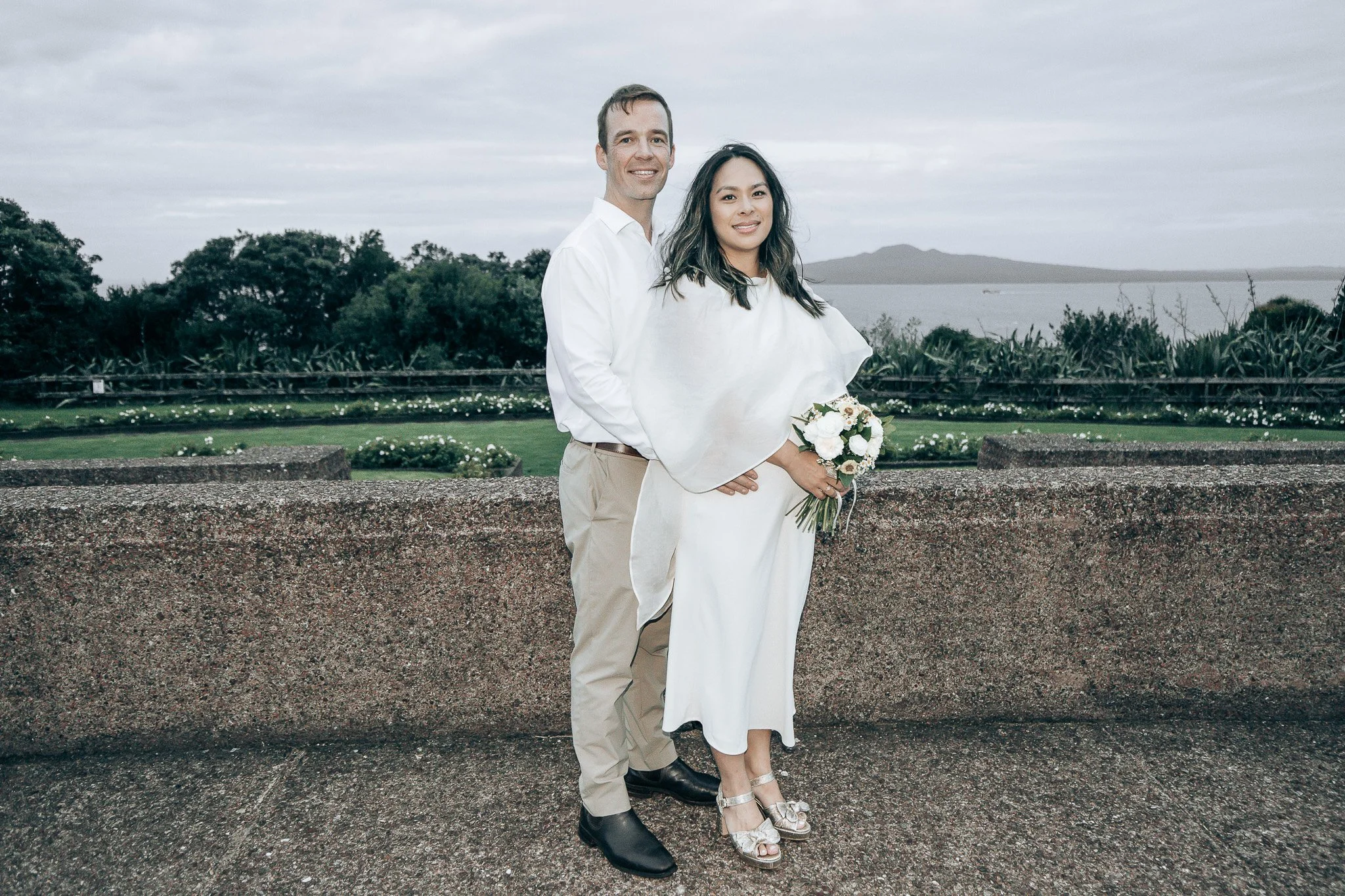A couple standing outdoors by a stone railing, with a scenic background including trees, a body of water, and distant hills or mountains. The man is wearing a white shirt and beige pants, and the woman is dressed in a white dress and holding a small 