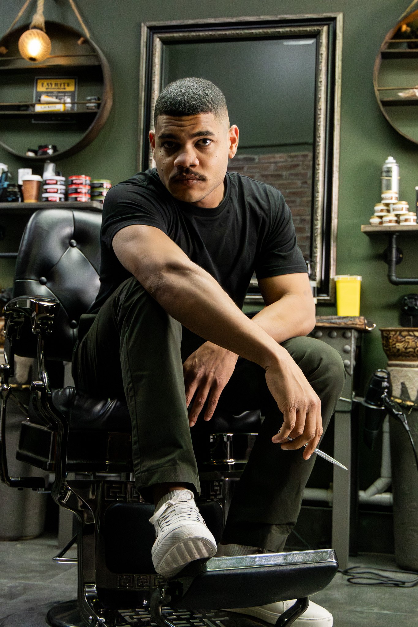 A young man with short hair and a mustache sitting in a barber's chair, looking directly at the camera with a serious expression, in a barbershop setting with shelves of hair products and a mirror in the background.