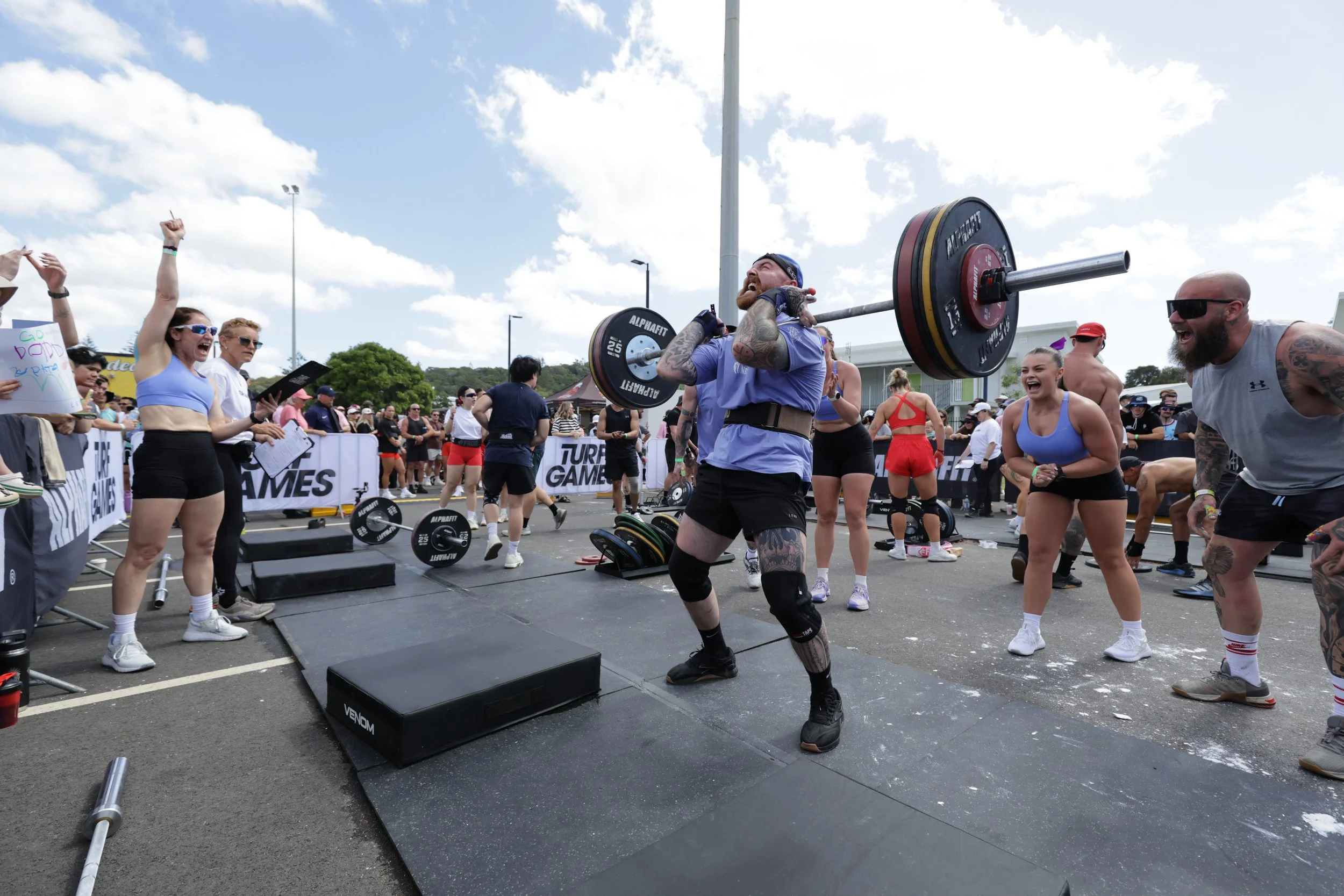 A man in athletic gear lifting a heavy barbell over his shoulders during a fitness competition or event, surrounded by a cheering crowd and other participants.
