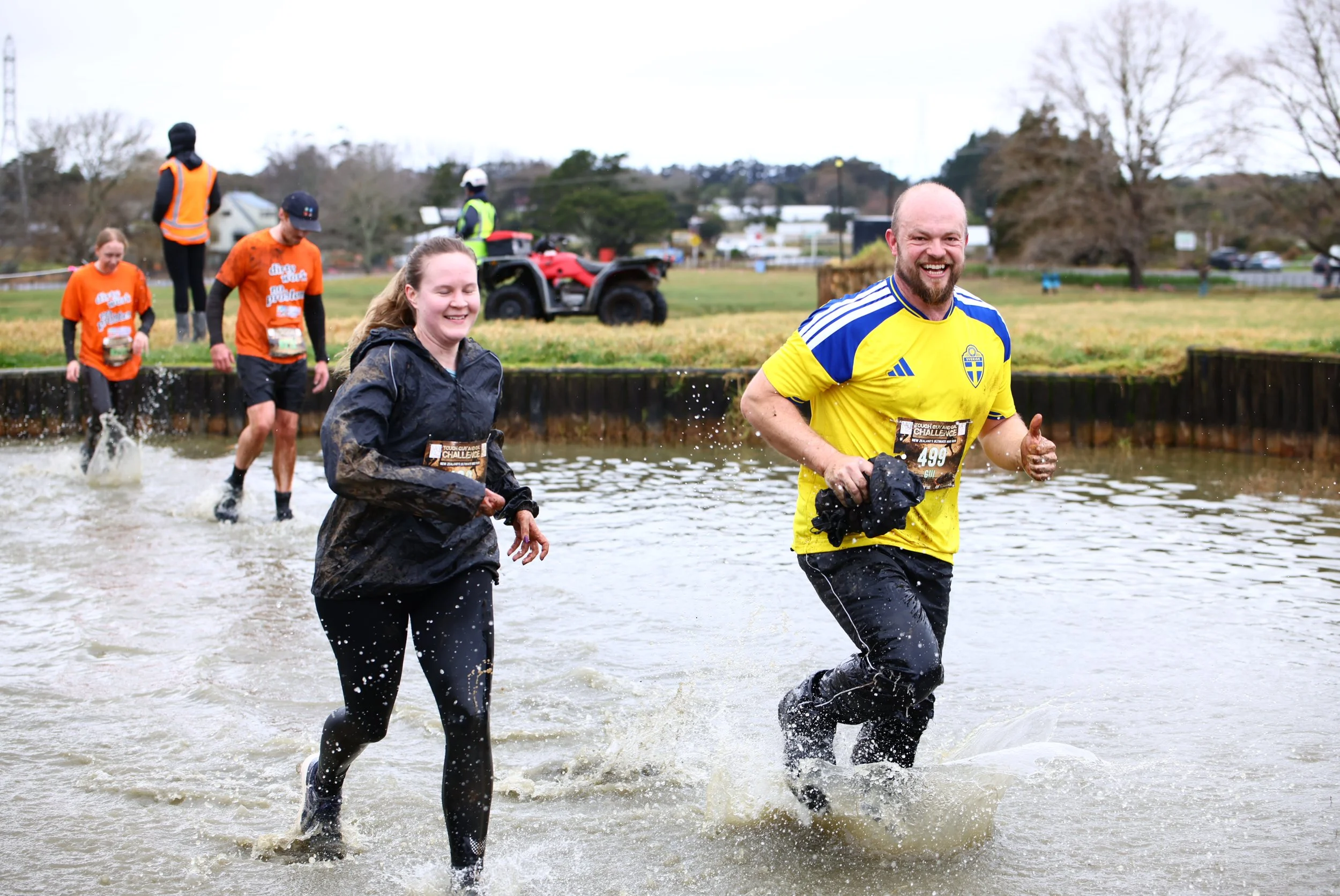 People running through a water obstacle during a challenge event outdoors, with trees and cloudy sky in the background.