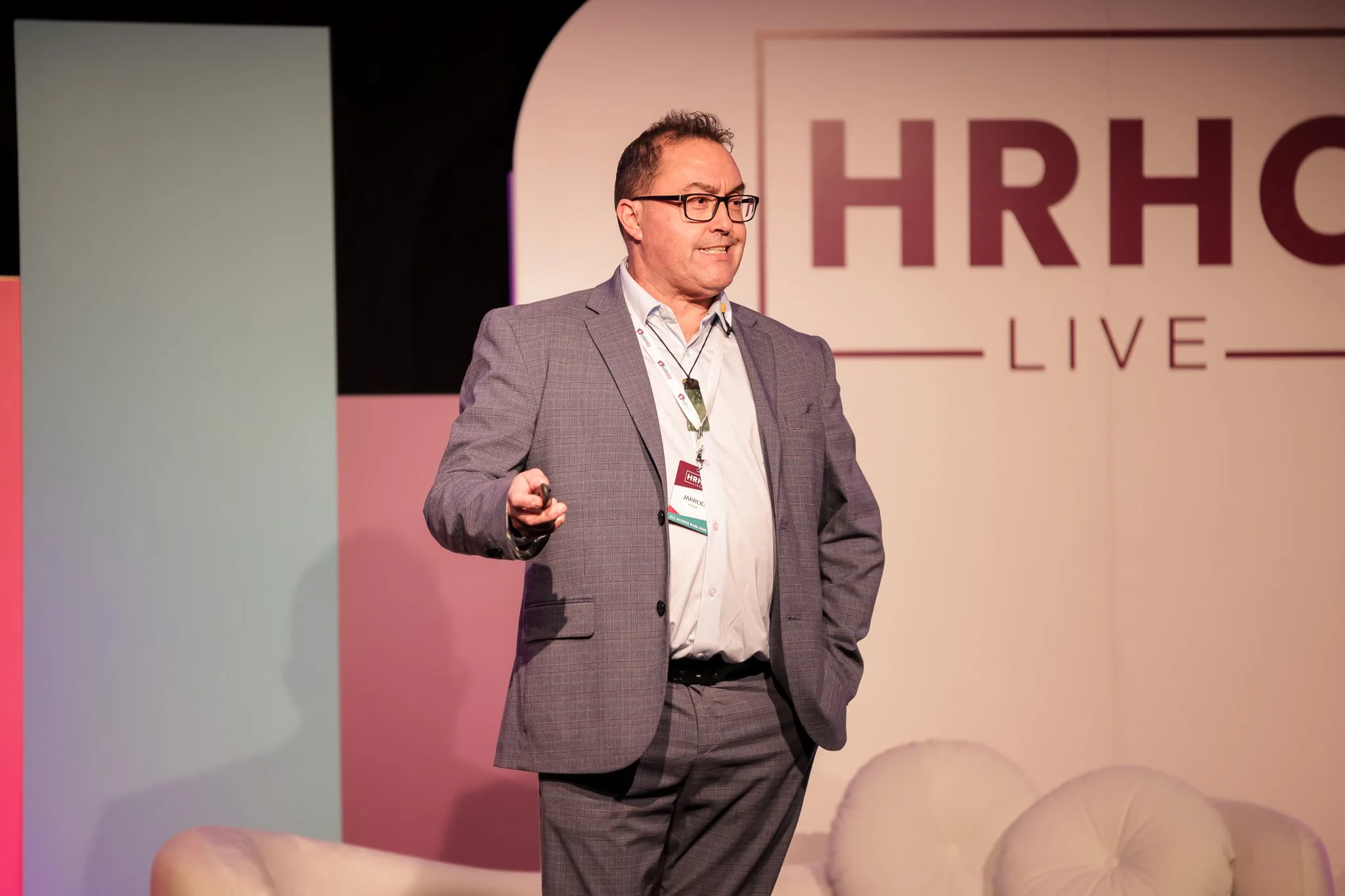 A man wearing glasses, a gray checkered suit, and a white shirt giving a presentation on a stage with a background that includes a large sign reading 'HR' and some white balloons.