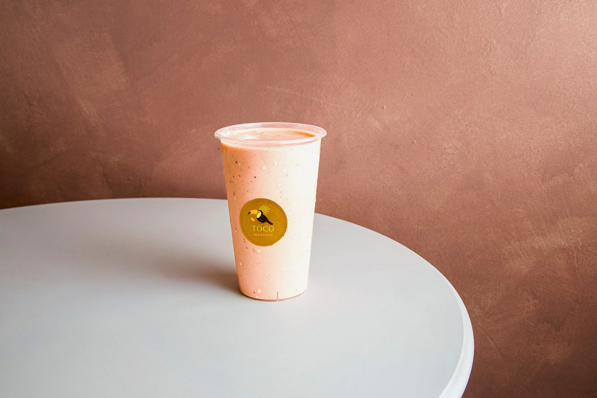 A plastic cup with a tropical bird logo labeled 'TOCO Açaí & Froyo,' filled with a pink and white blended frozen treat, placed on a white surface against a brown textured background.