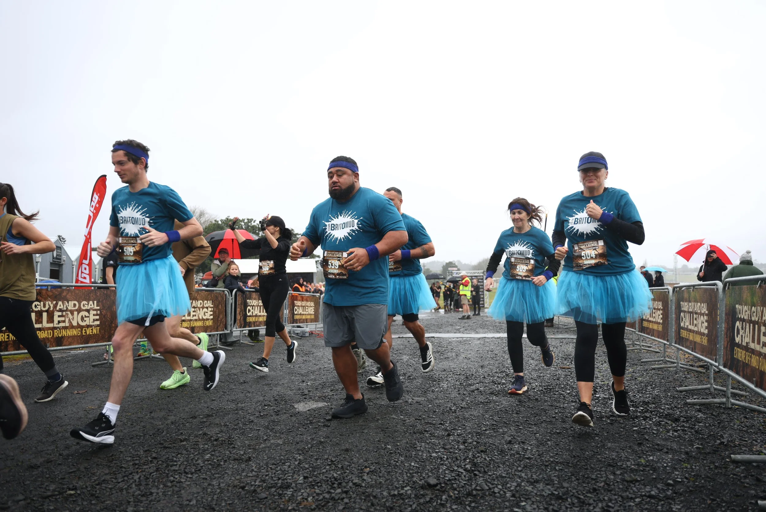 People participating in a race wearing blue t-shirts and tutus, running outdoors on a cloudy day with spectators and event banners in the background.