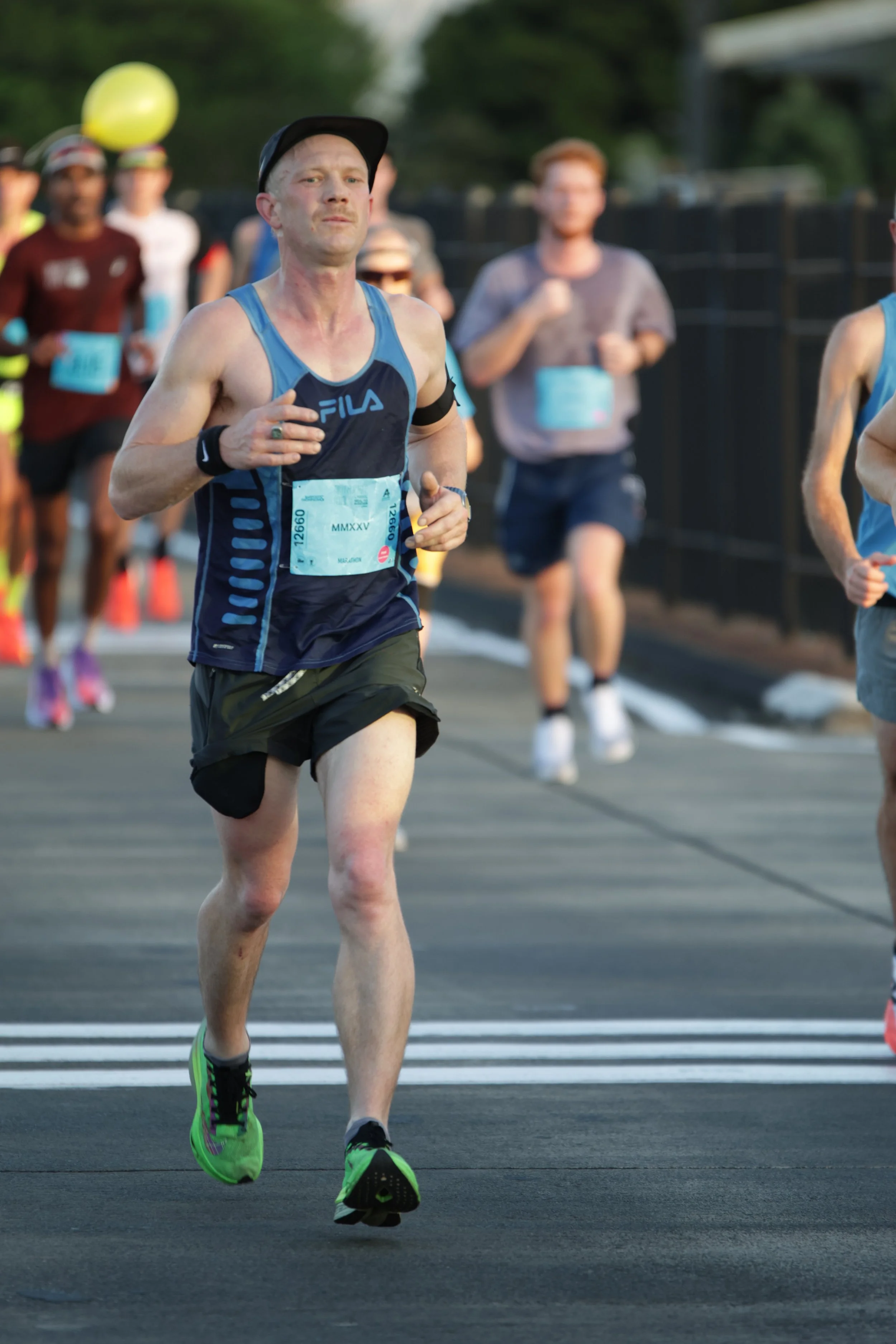 A male runner in a blue FILA tank top and black shorts participating in a race, with other runners behind him on a road marked with a white line, possibly during early morning or late afternoon.