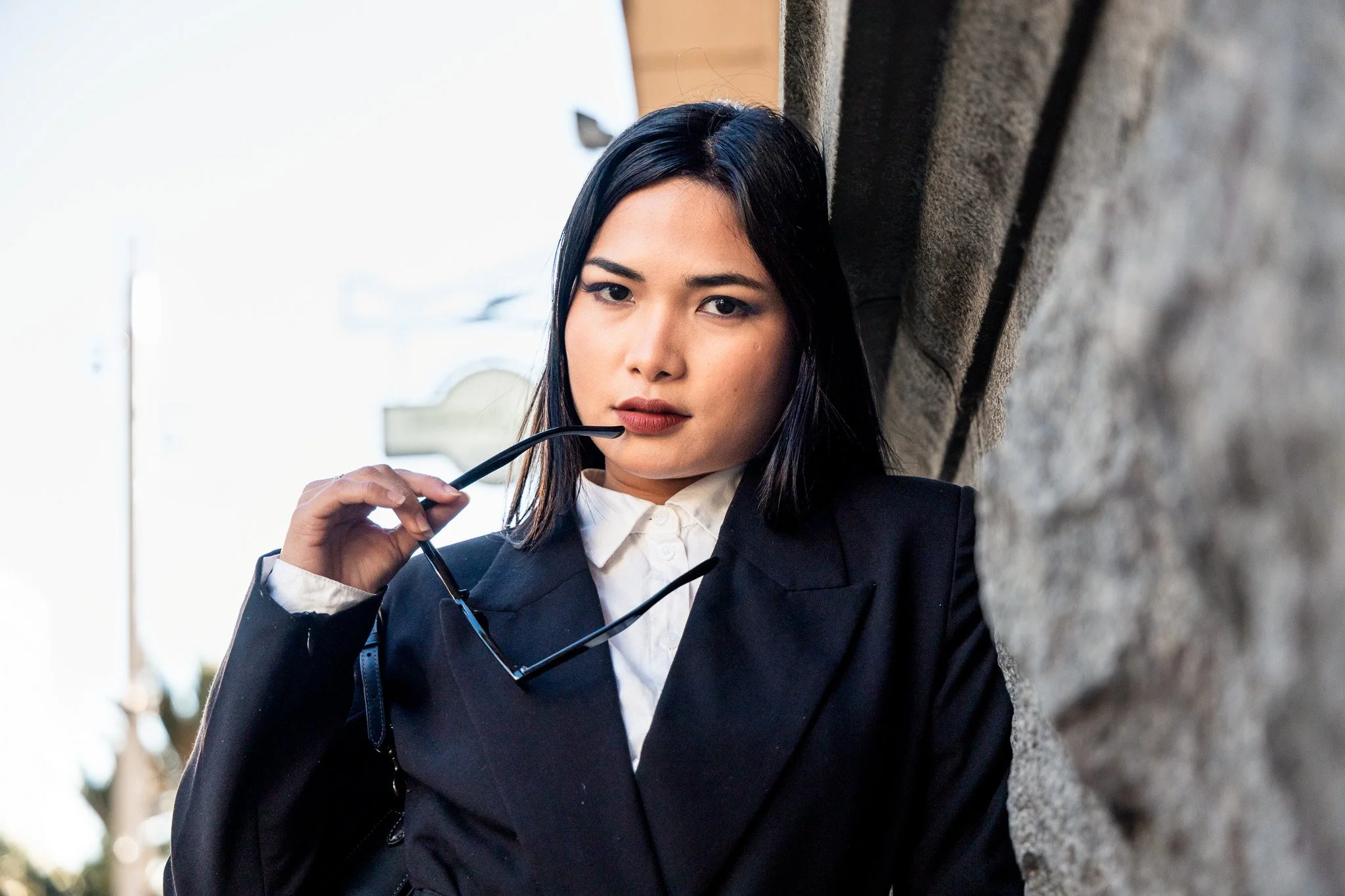 Portrait of a woman with black hair, wearing a black blazer and white shirt, standing outdoors against a stone wall, holding glasses near her mouth.