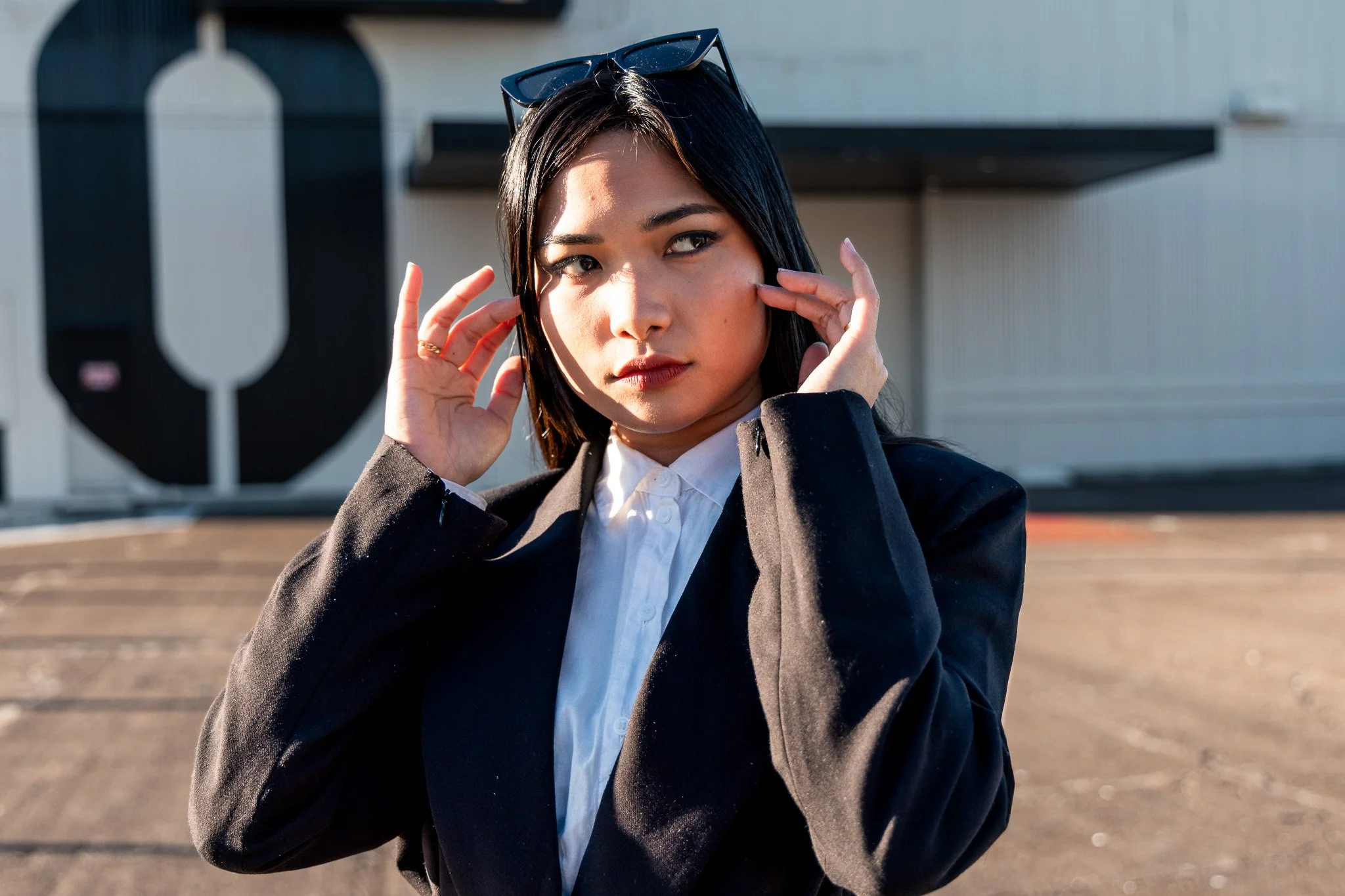 A young woman with black hair and sunglasses on her head, wearing a white shirt and black blazer, looks at the camera outdoors on a sunny day.
