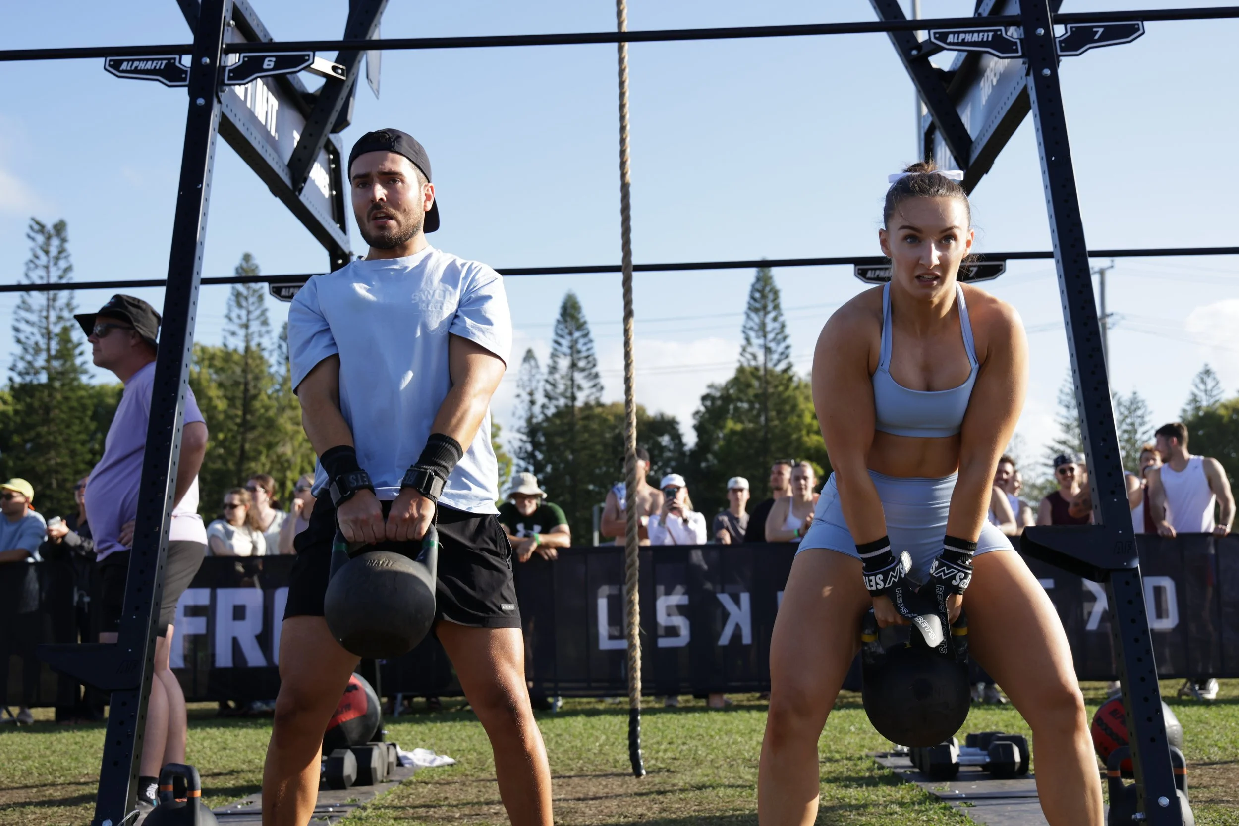 Two individuals participate in an outdoor fitness event. The man on the left is lifting a kettlebell, and the woman on the right is holding a sled while wearing workout gloves. They are surrounded by a crowd of spectators under a clear sky.