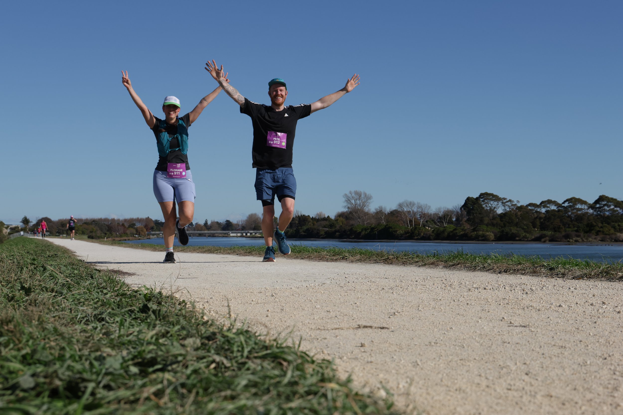 A man and woman running on a sidewalk beside a body of water, both smiling with their arms raised, against a clear blue sky.