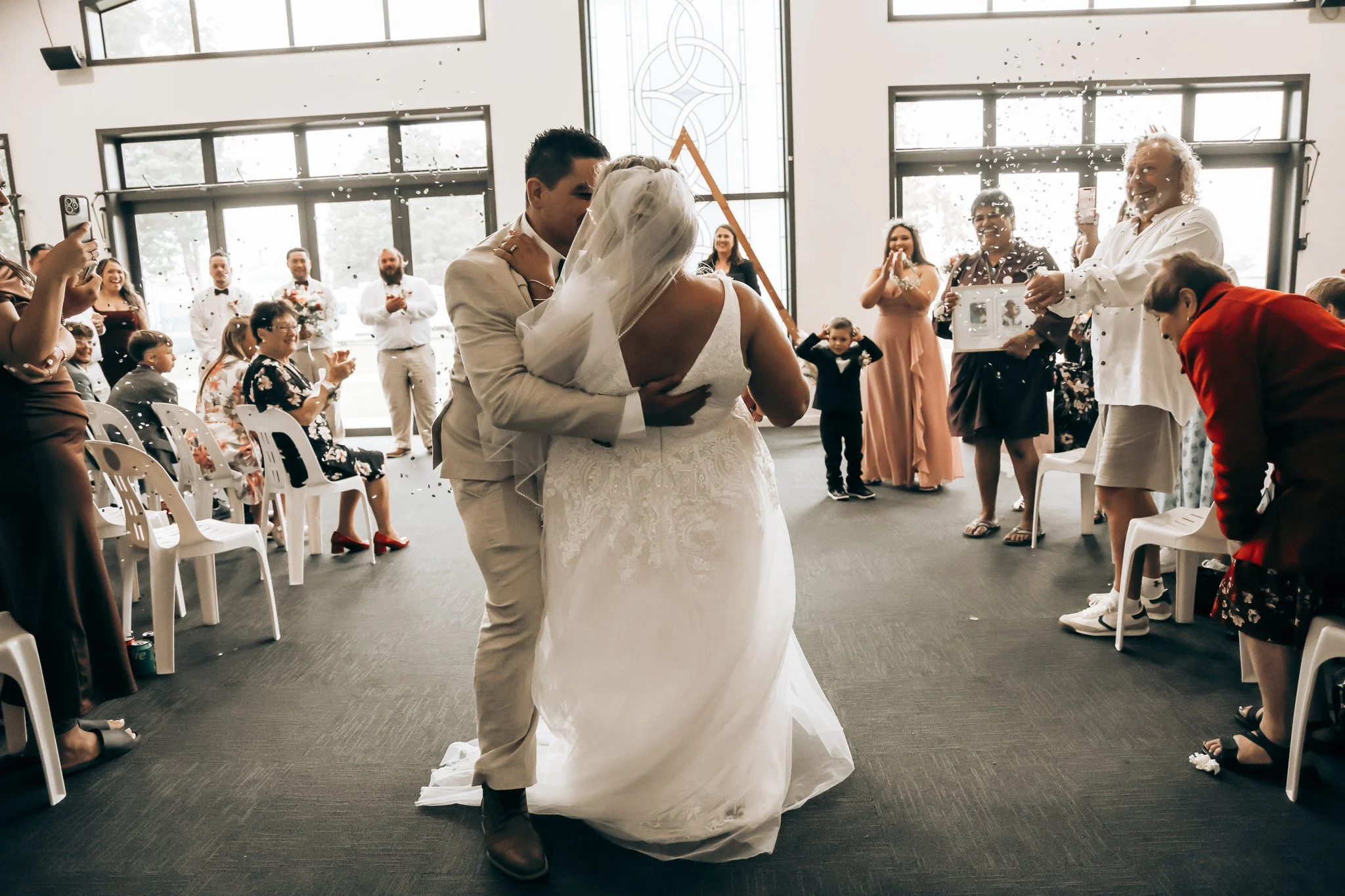 A bride and groom are sharing a dance in a brightly lit wedding reception hall, while guests are clapping and taking photos around them.