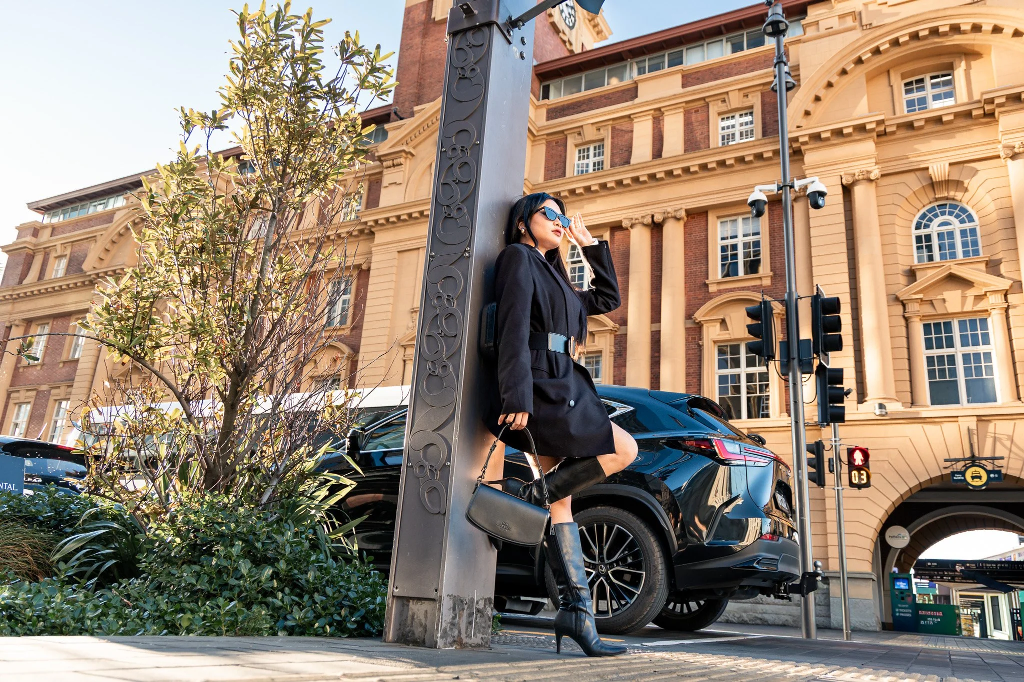 A woman wearing sunglasses and a black coat, leaning against a lamppost on a city street. She is holding her hand near her face, with a large building with ornate architecture in the background. There are cars parked nearby and a traffic signal showi