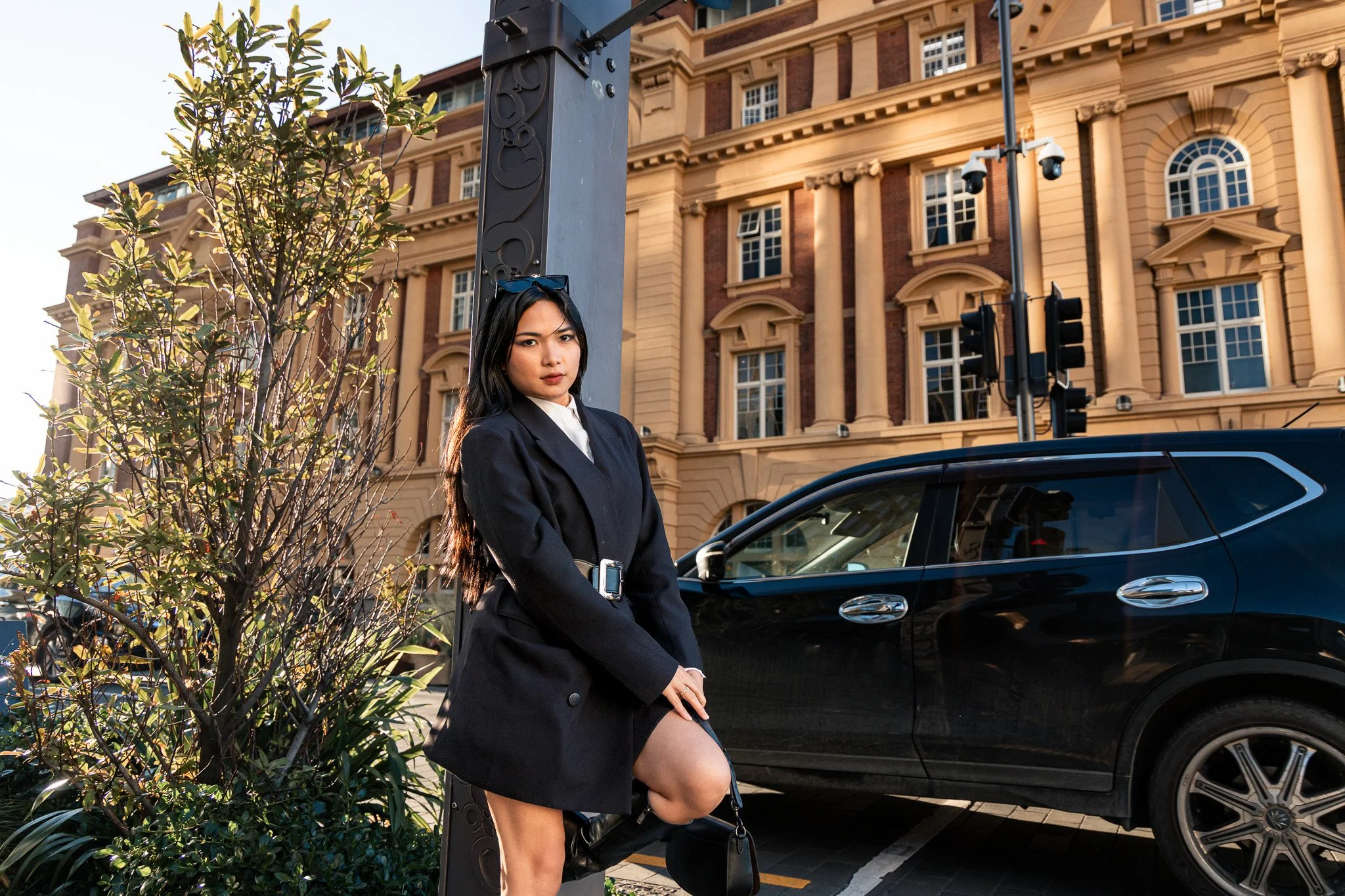 A woman in a black blazer and white shirt sitting on a ledge at a city street corner with a multi-story brick and beige building in the background, a black car parked nearby, and a police surveillance camera mounted on a pole with sunglasses resting 