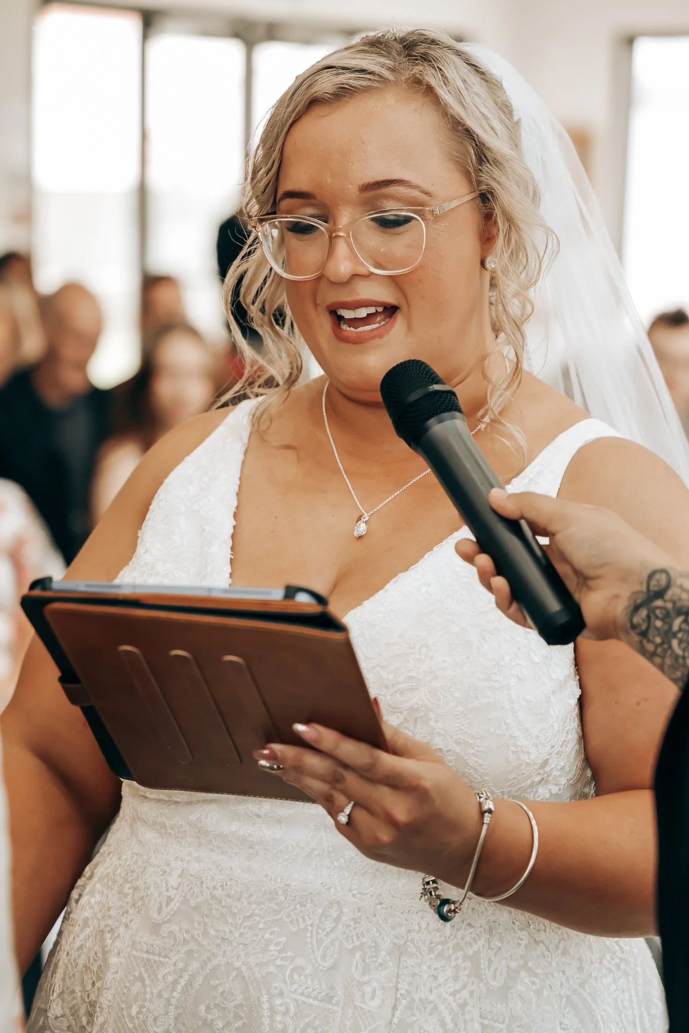 A woman wearing glasses, a white wedding dress, and veil reading vows from a book during a wedding ceremony. An individual is holding a microphone close to her mouth. Guests are blurred in the background.