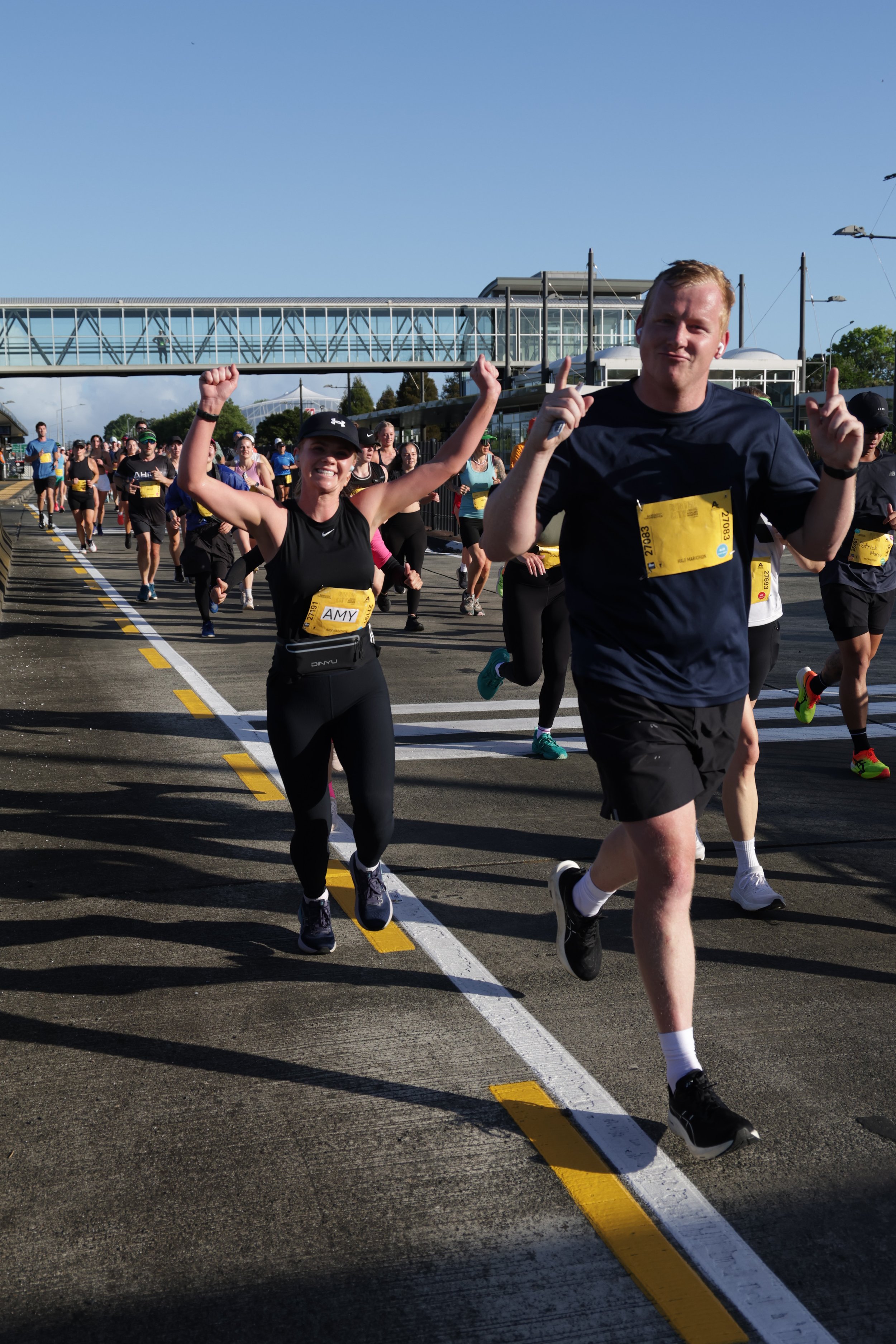 A group of marathon runners participating in a race, with ablu sky and a modern building with a pedestrian bridge in the background.