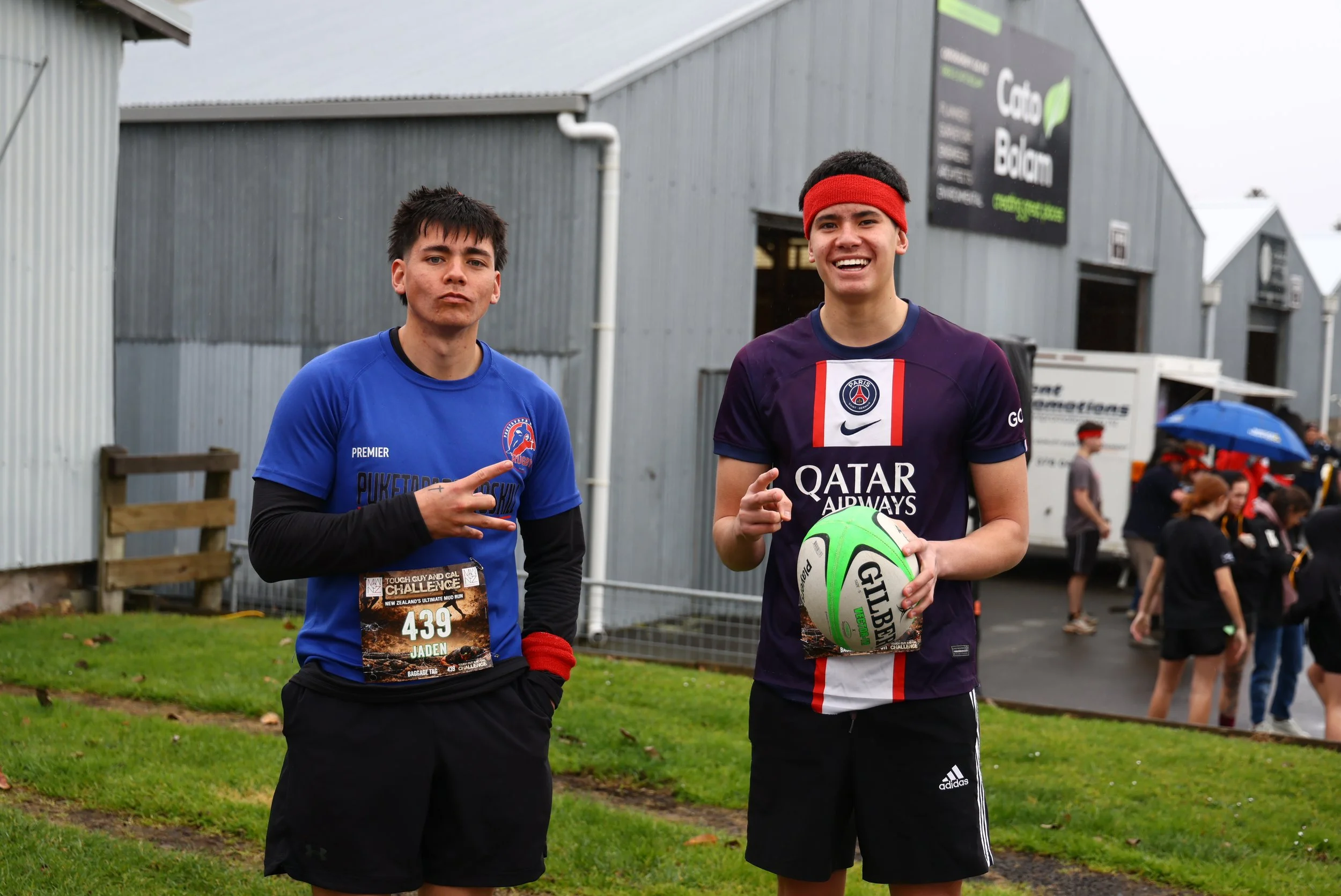 Two young men standing outdoors, one holding a soccer ball and both smiling, at a sporting event with other people and a gray building in the background.