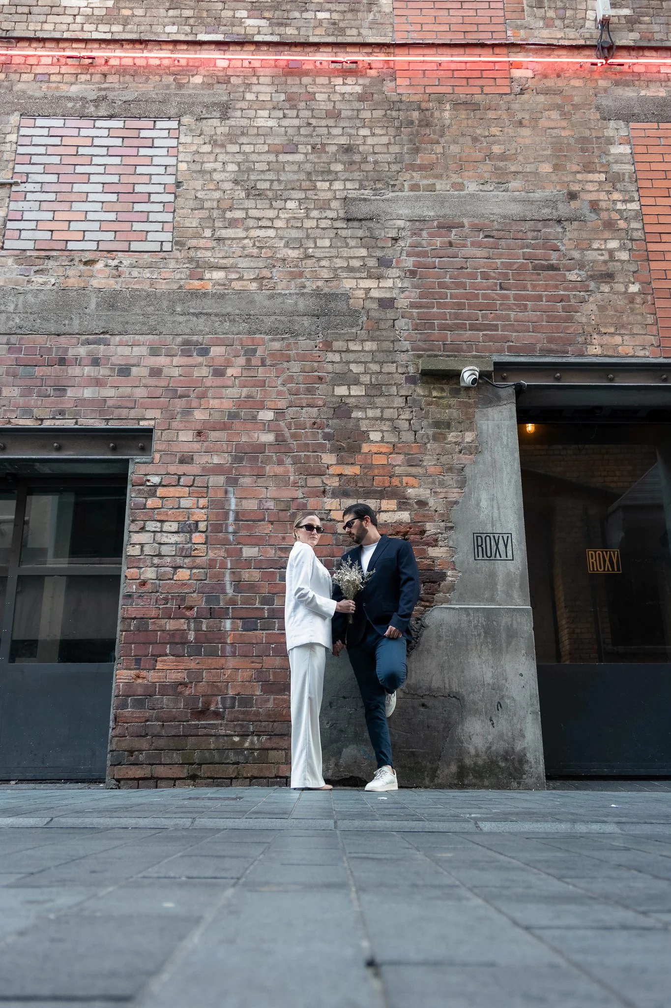 Two people, a woman in a white suit and a man in a dark jacket, stand close together in front of a brick wall, with the woman holding a bouquet of flowers, outside a building labeled 'ROXY'.