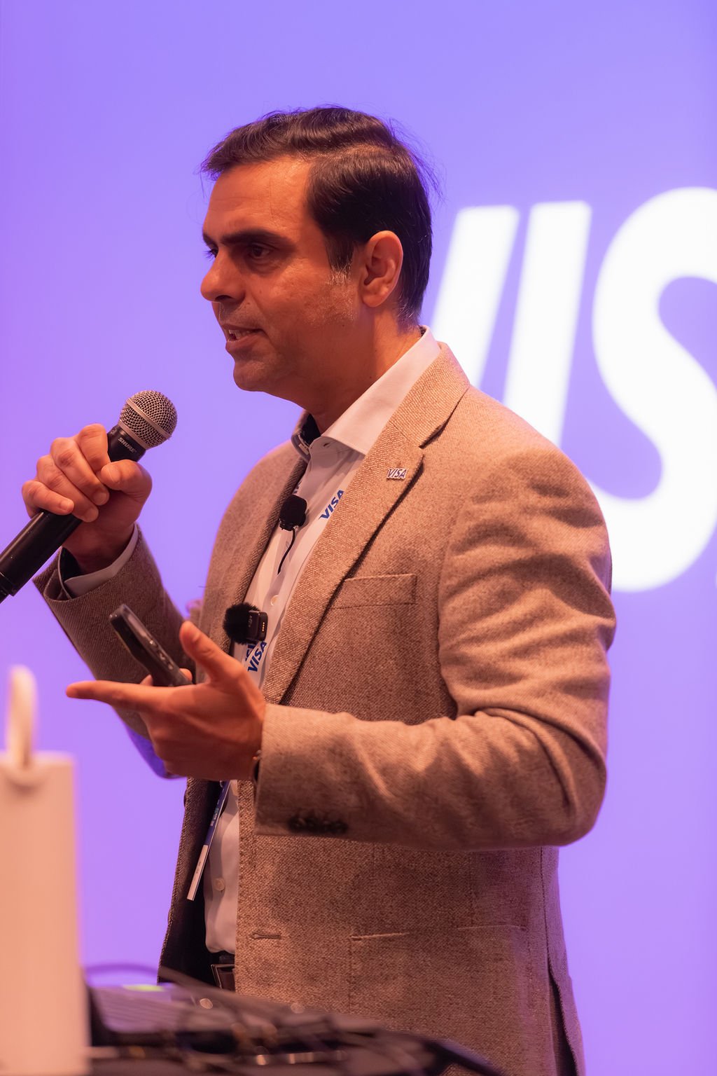 A man in a beige blazer and white shirt speaking into a microphone during a presentation with a blurred purple background displaying the partial 'VISA' logo.