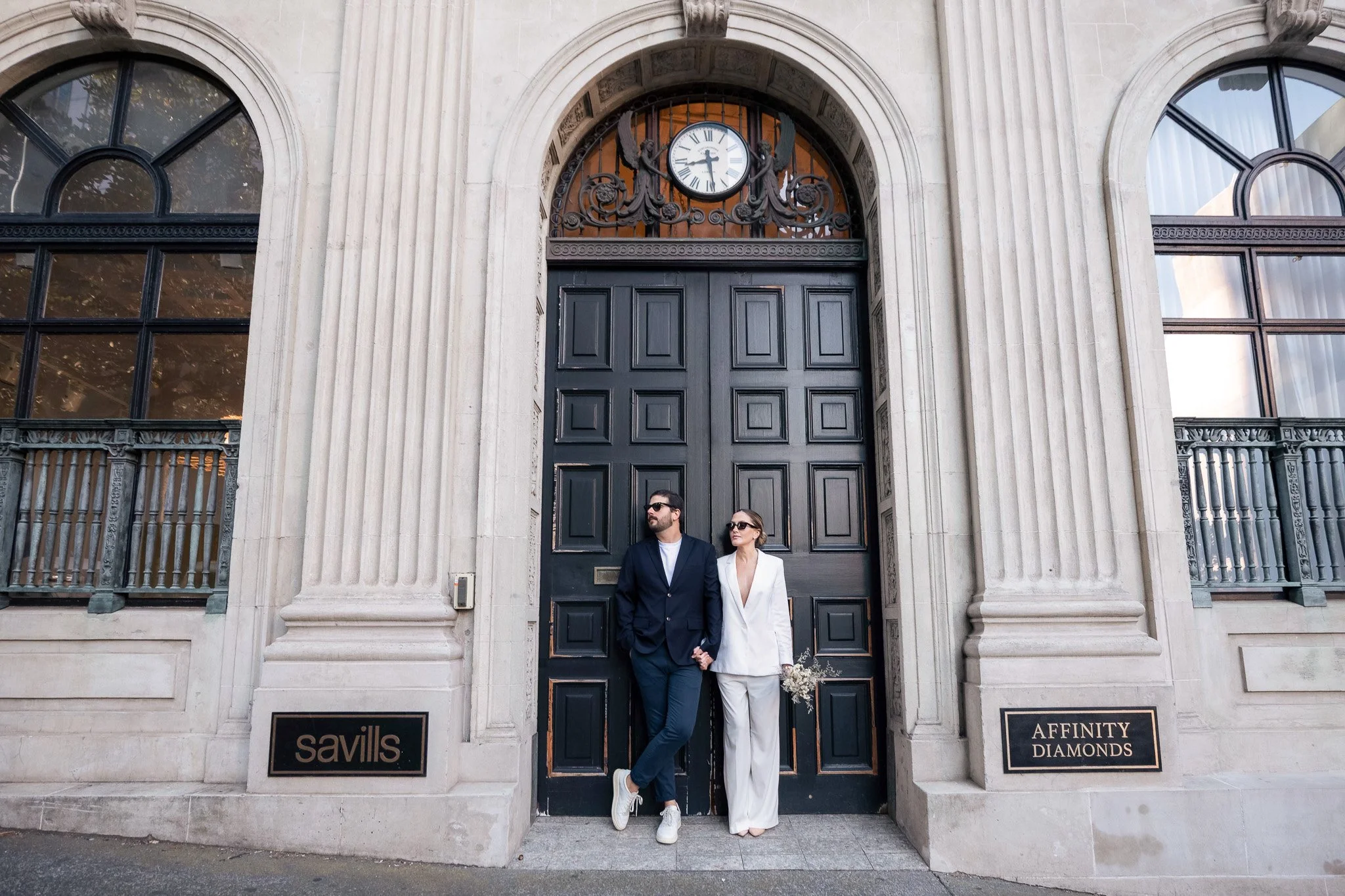 A man and woman in formal attire standing together outside a large building with a clock above the entrance, black door, and signage for Savills and Affinity Diamonds.