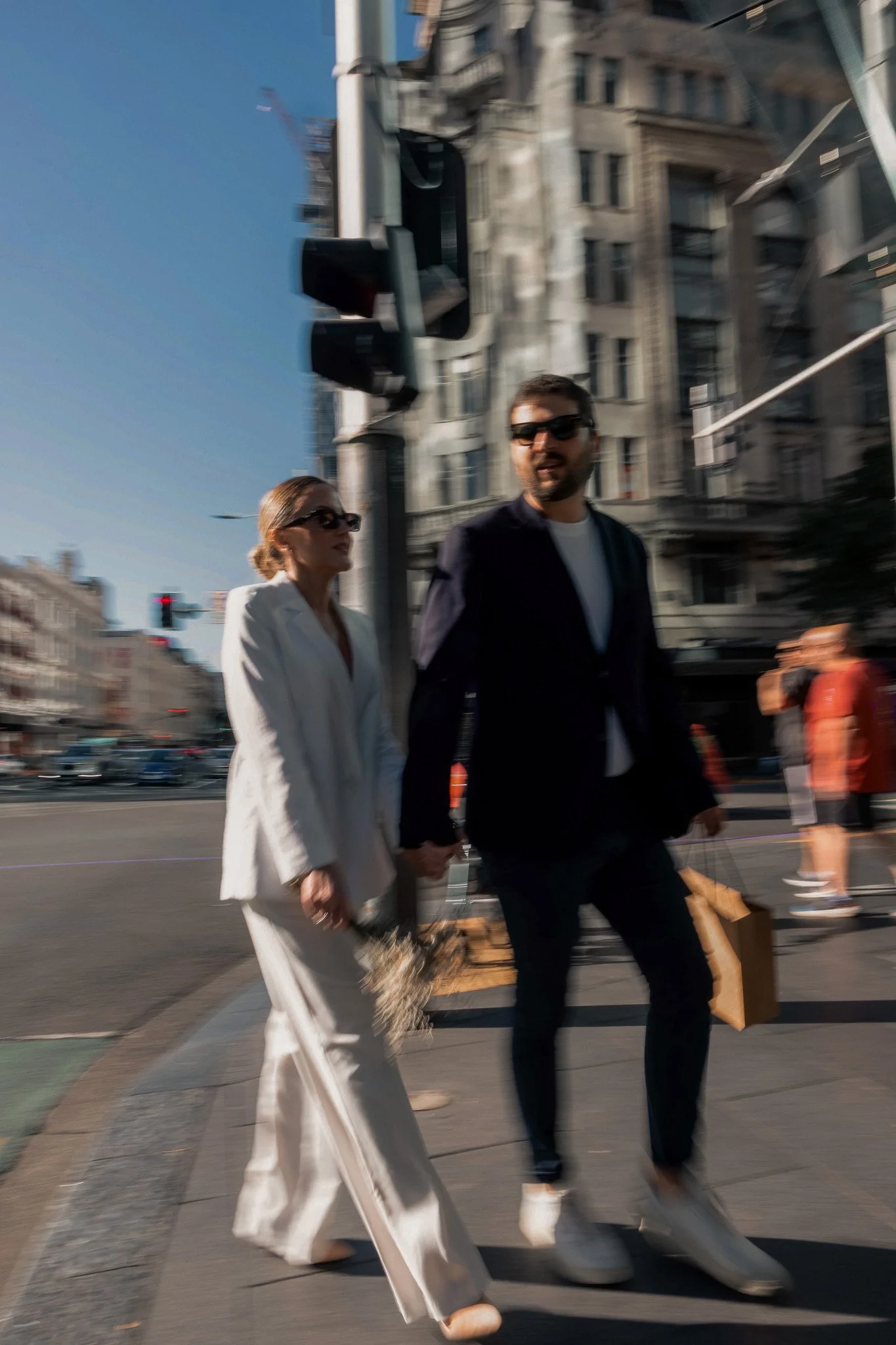 Two people, a woman dressed in a white suit and a man in dark clothing, walking in a city street with tall buildings, traffic lights, and a clear blue sky, holding hands and carrying shopping bags.