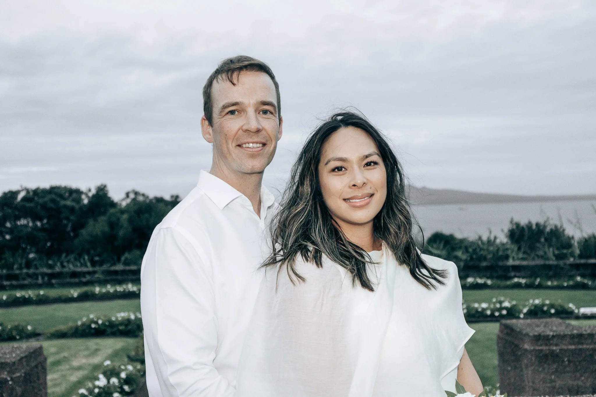 A happy couple standing outdoors in front of a body of water, with trees and cloudy sky in the background. The man is wearing a white shirt with short brown hair, and the woman has long wavy dark hair with highlights, wearing a white top.