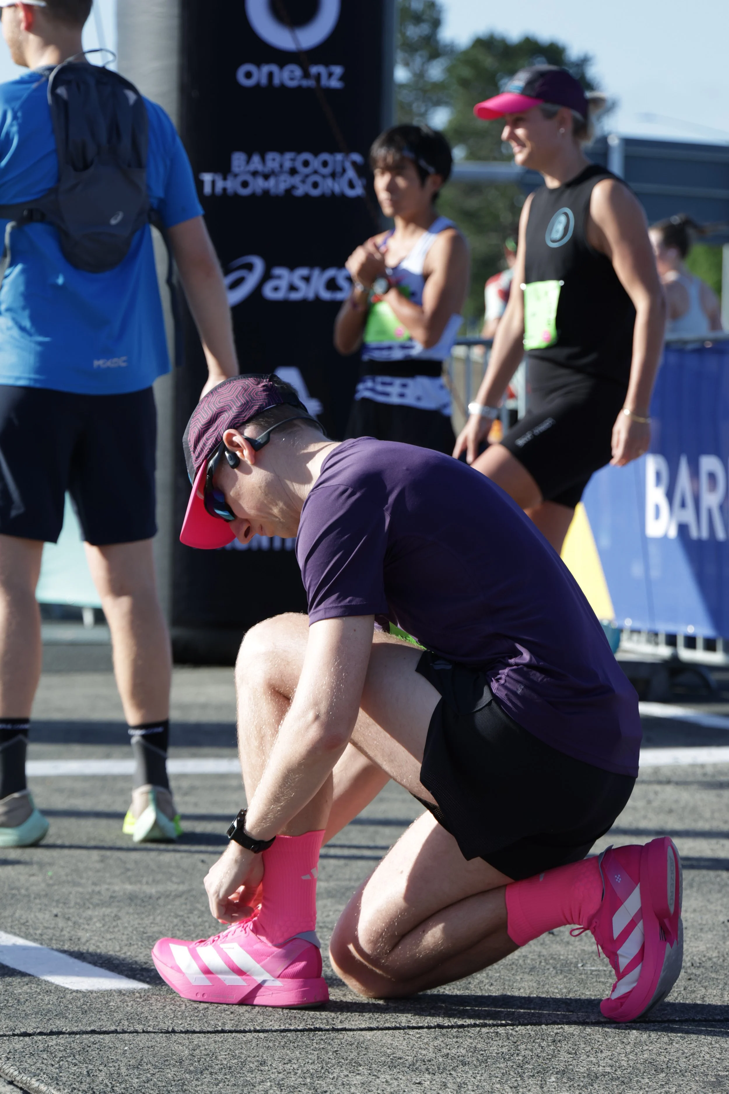 A person wearing a purple shirt, black shorts, pink socks, and pink running shoes is kneeling on one knee, tying their shoelace during a race or athletic event. Other participants are visible in the background, along with banners and a starting line 