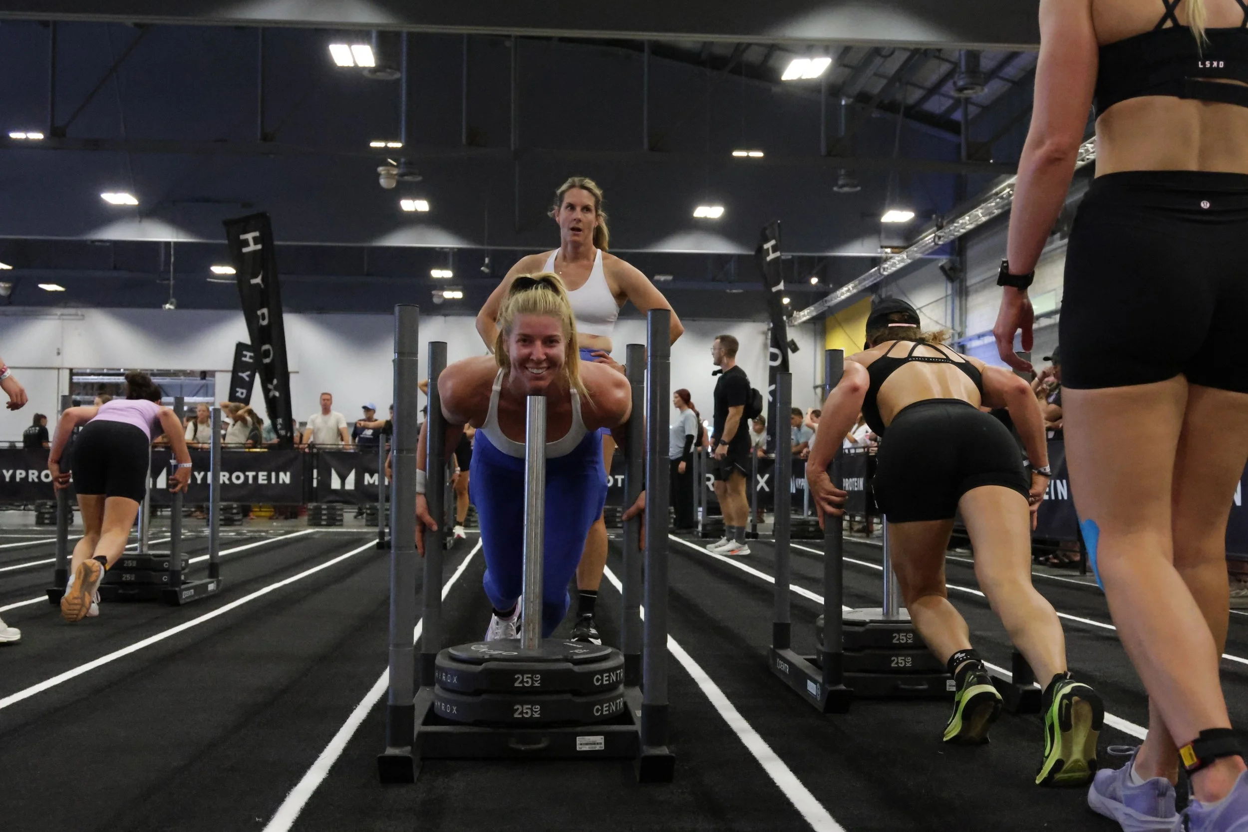 A woman in athletic clothes is performing a plank exercise on a stack of weight plates at a busy indoor gym. Other people are also exercising in the background.