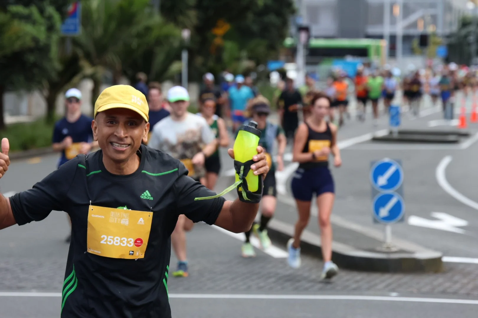 Smiling male runner with yellow cap and water bottle participating in a marathon, with several other runners in the background on a city street.