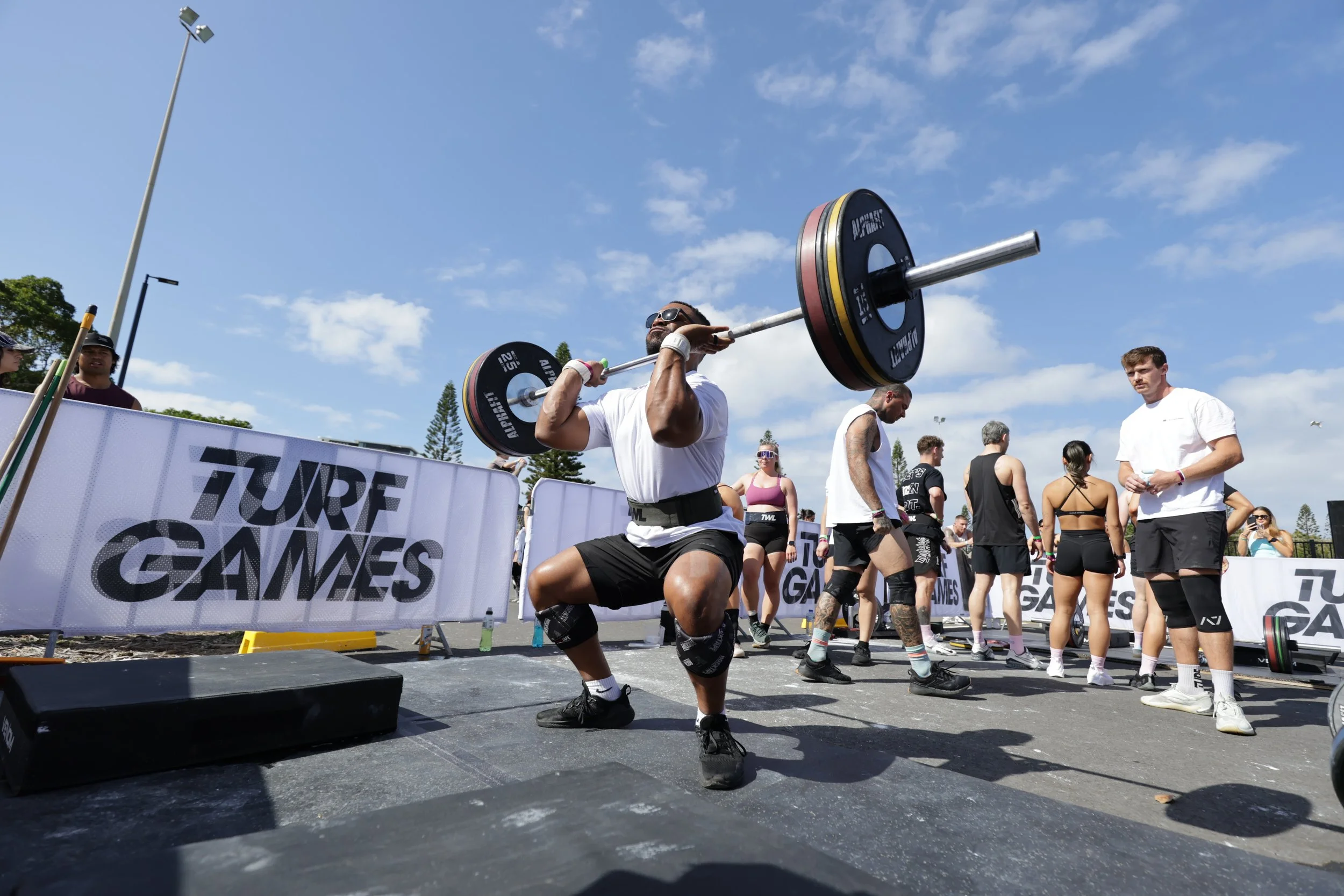 A man lifting a barbell with weights during a weightlifting event at an outdoor competition, with spectators and other athletes watching.
