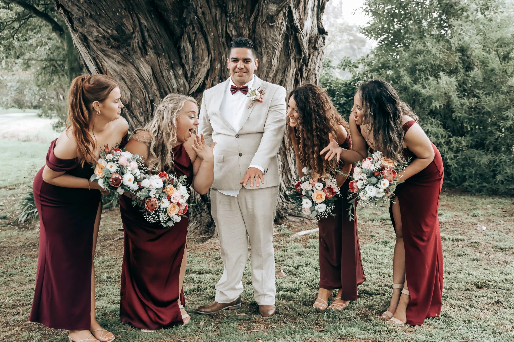 A man in a white suit with a bow tie and boutonniere stands under a tree, surrounded by four women in burgundy dresses holding bouquets, all smiling and engaging in conversation outdoors during daytime.
