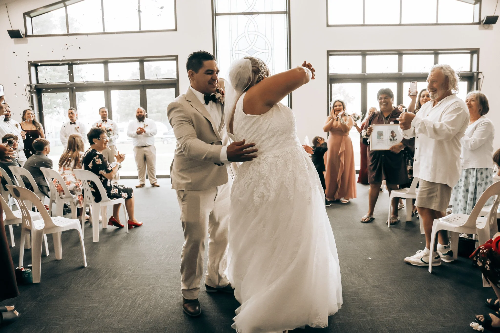 A wedding ceremony inside a church or hall with large windows, showing a bride and groom dancing while surrounded by smiling guests, some taking photos.