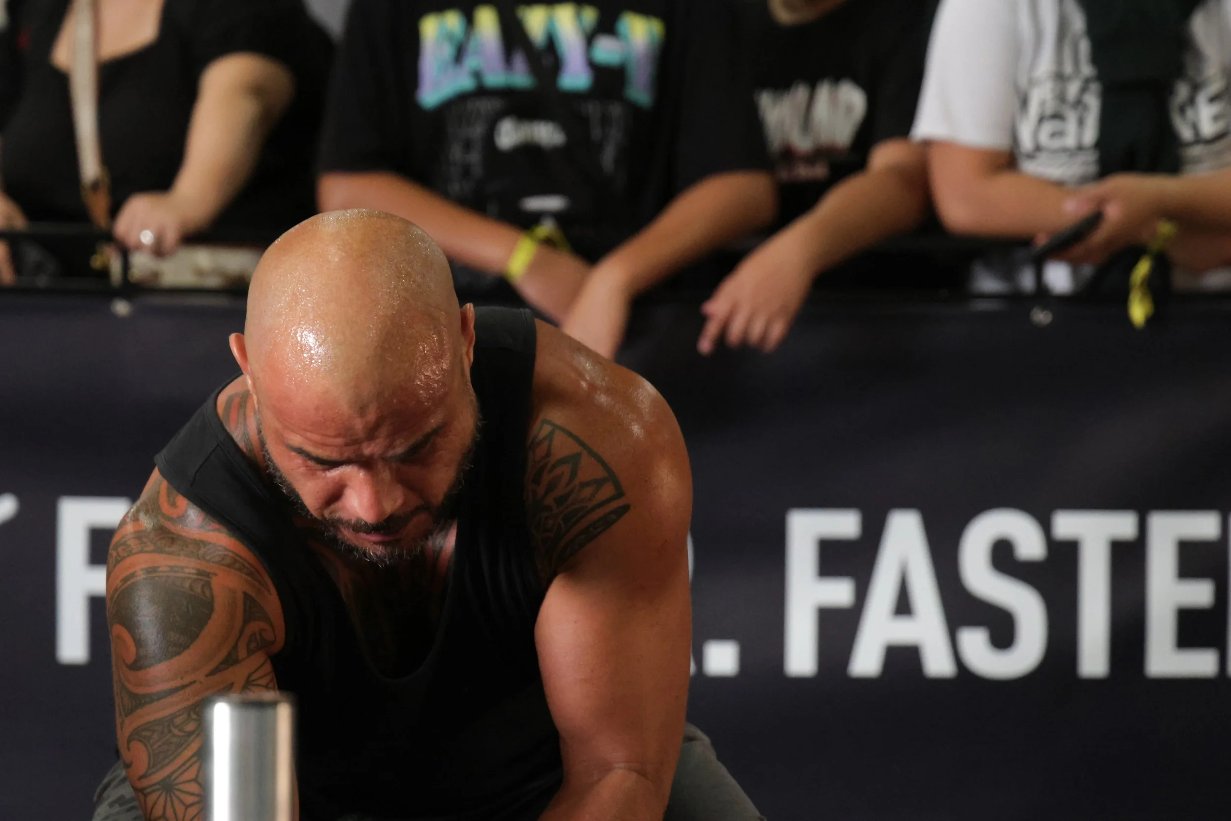 A man with tattoos is intensely focused during a workout, with others in the background seated and using their phones at a fitness event.
