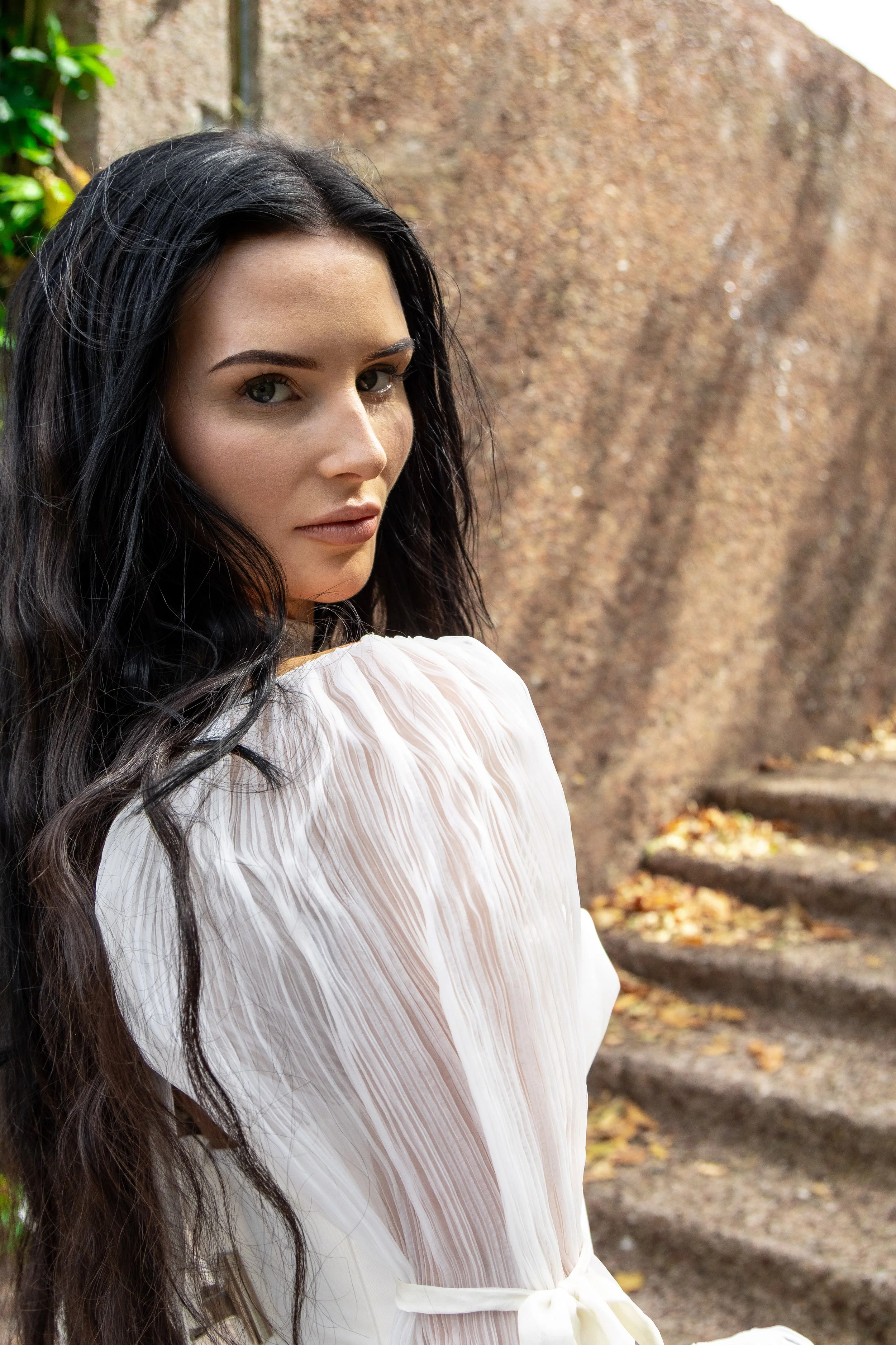 A woman with long dark hair and blue eyes, wearing a white, pleated dress with a bow at the waist, standing outdoors near a large rock with fallen autumn leaves on the ground.