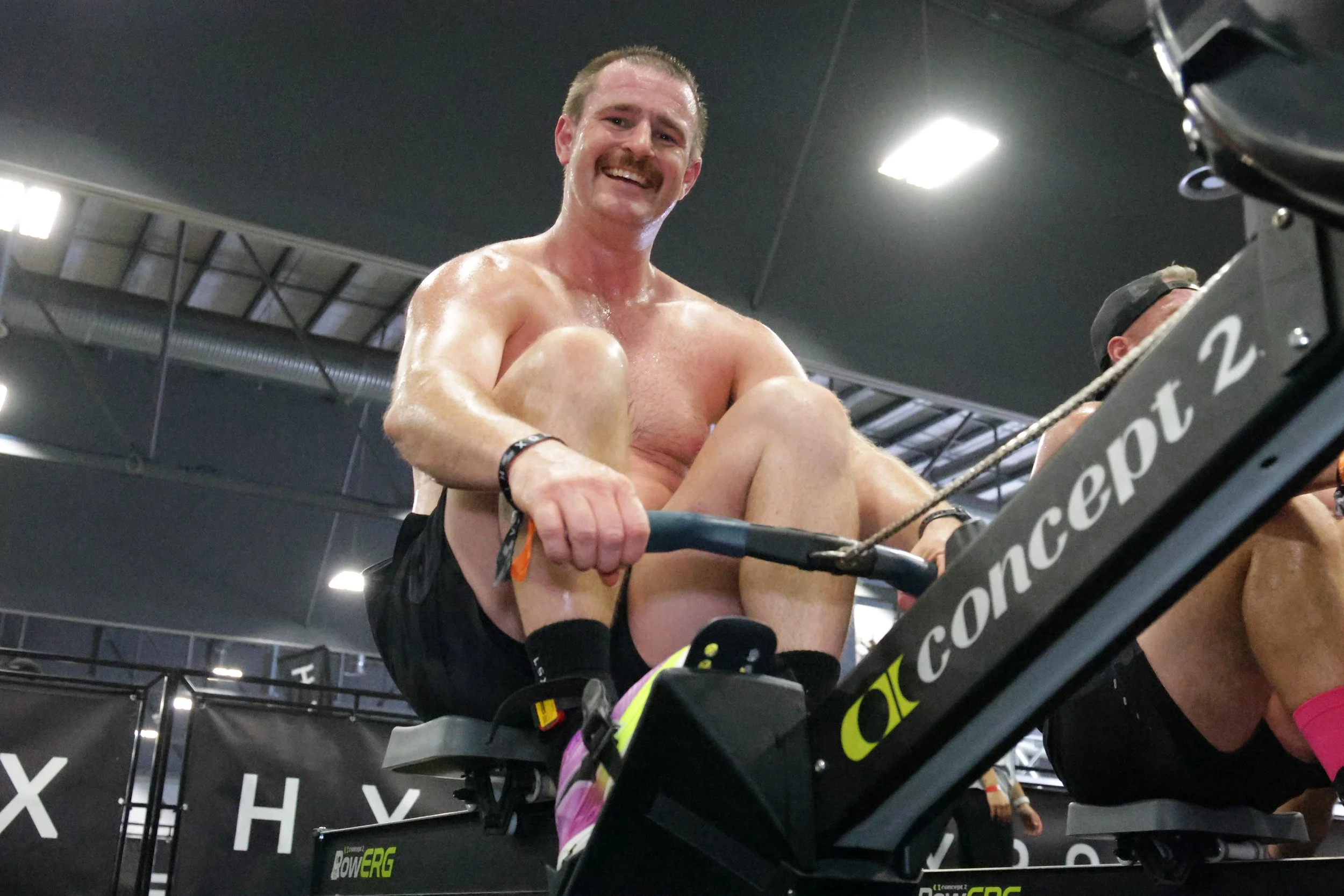 A shirtless man with a mustache and a black wristband, smiling and sitting in a rowing machine at a gym.