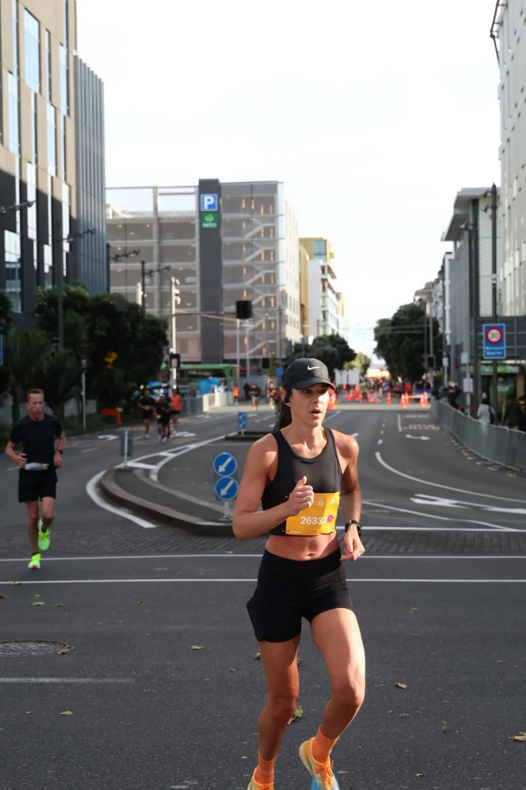 A woman running in a city marathon, wearing a black sports outfit, a black cap, and orange running shoes, with other runners and city buildings in the background.