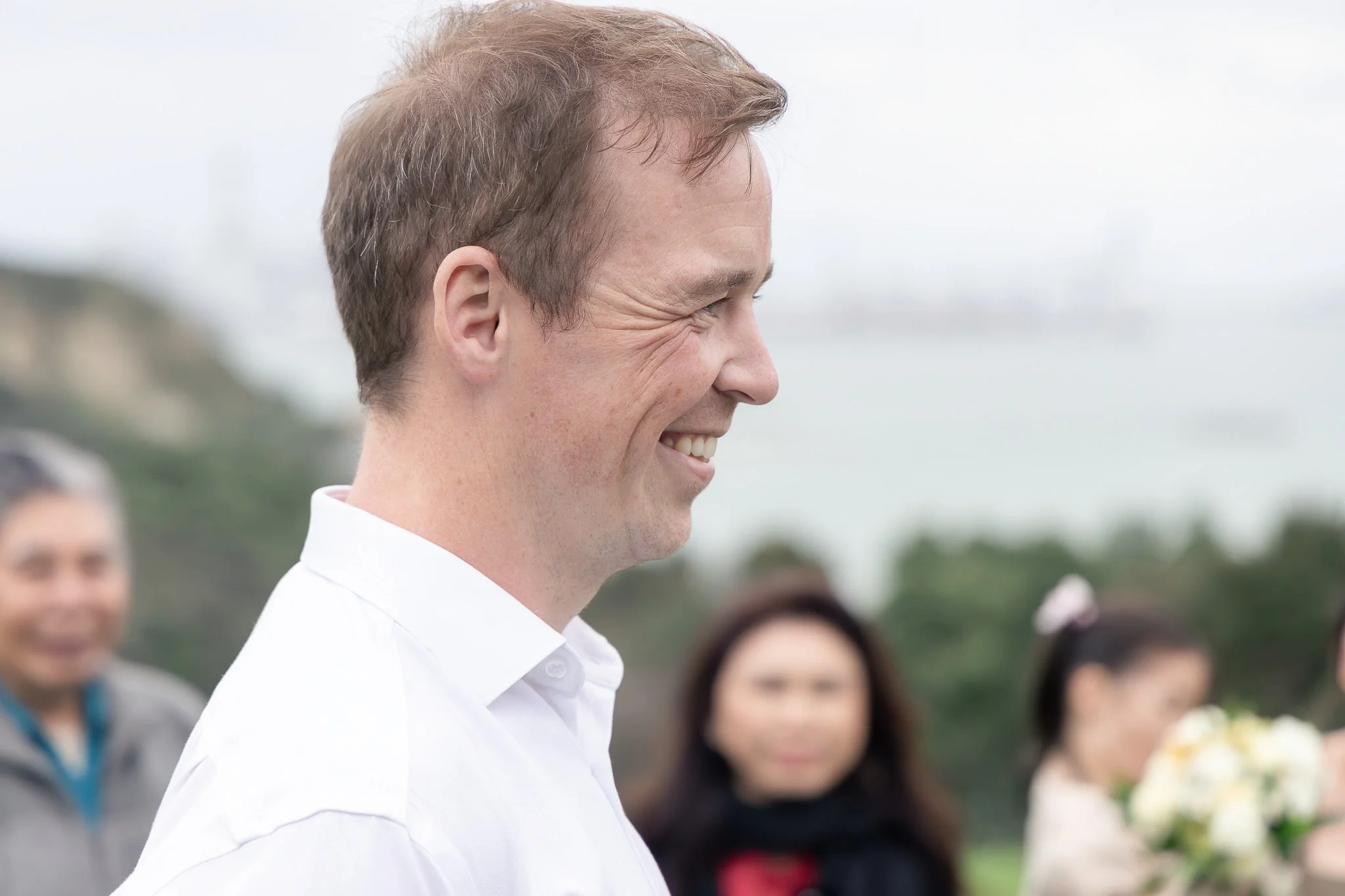 Side profile of a smiling man in a white shirt outdoors with a cloudy sky, blurred crowd in the background.