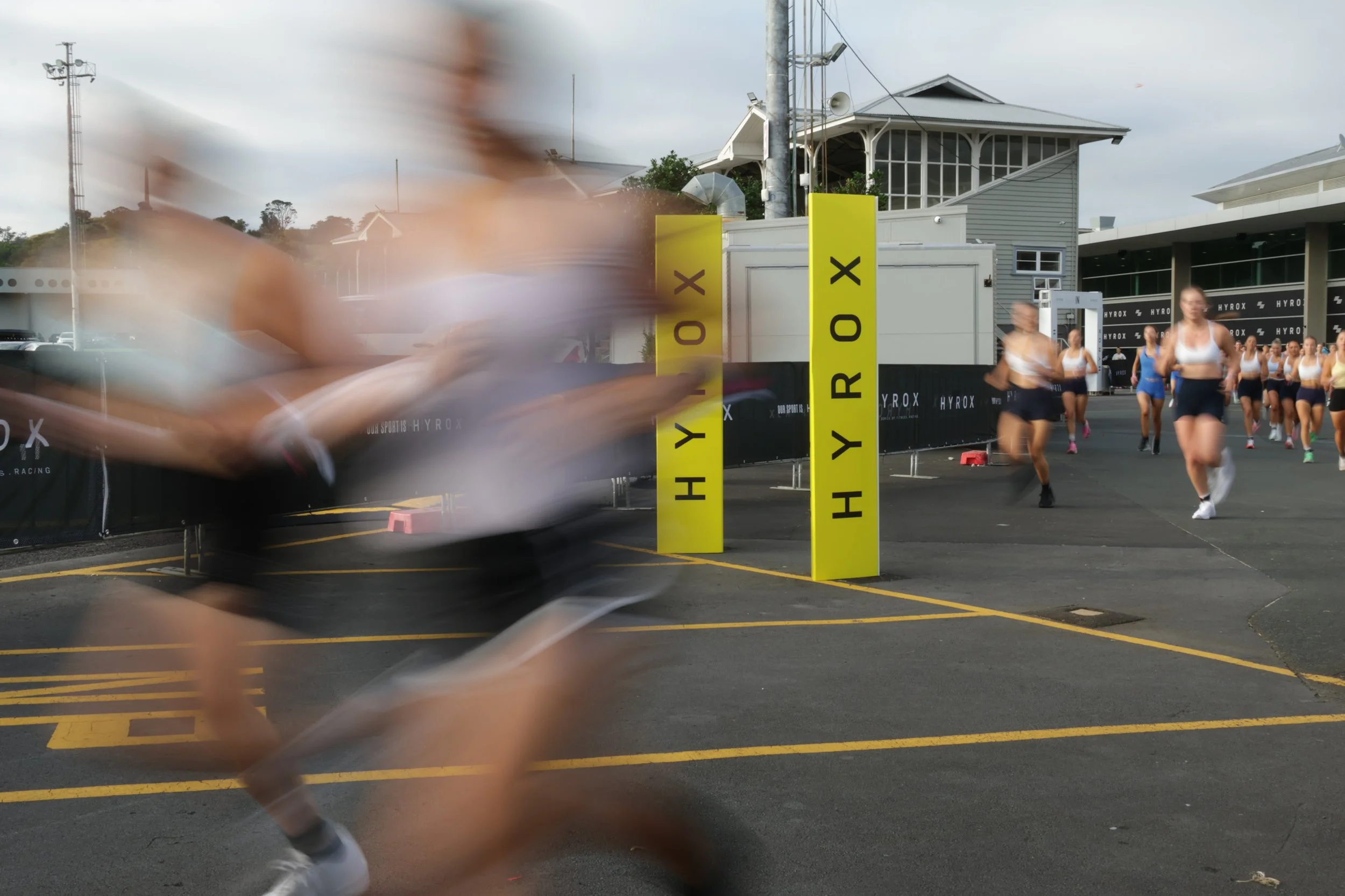 Blurred image of a group of women running in a race, with race signs and a building in the background, and a person running past in the foreground.