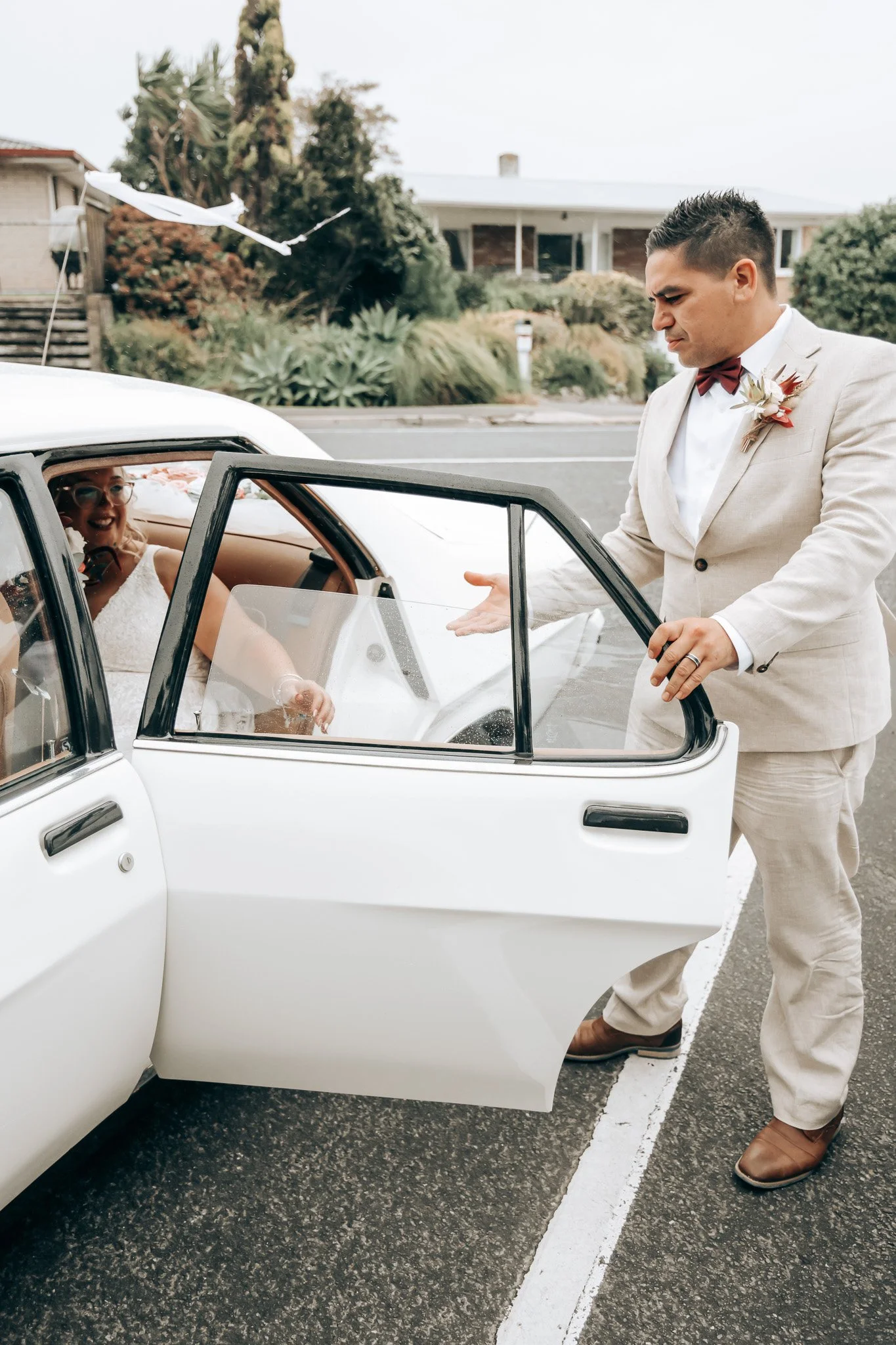 A man in a beige suit and bow tie standing outside next to a white vintage car with a woman sitting inside, smiling, through the open car door. The woman appears to be in bridal attire, and the man is gesturing towards her. In the background, there a