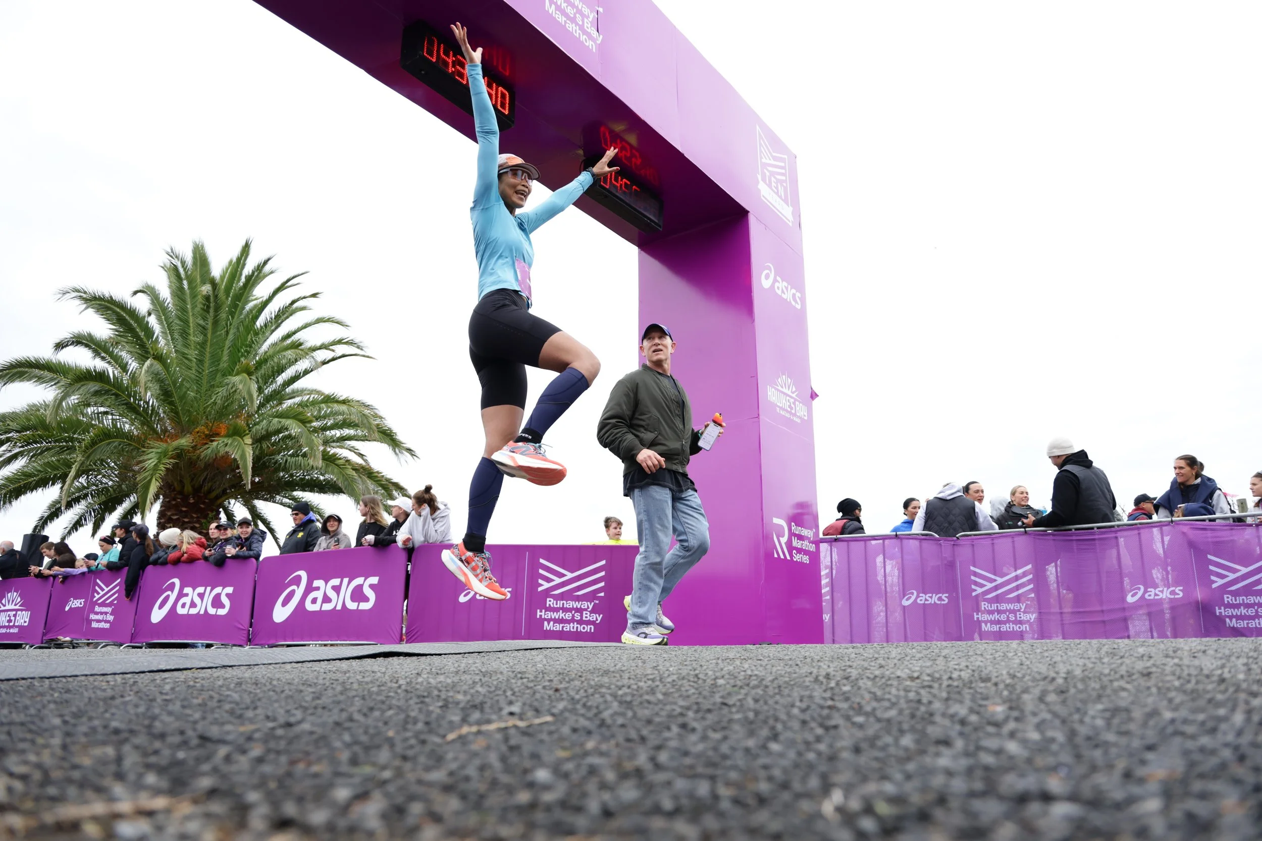 A woman in athletic clothing and sneakers crossing the finish line of a marathon with her arms raised in victory, while a man in a jacket and cap stands nearby. There are spectators behind a purple barrier, and palm trees are visible in the backgroun