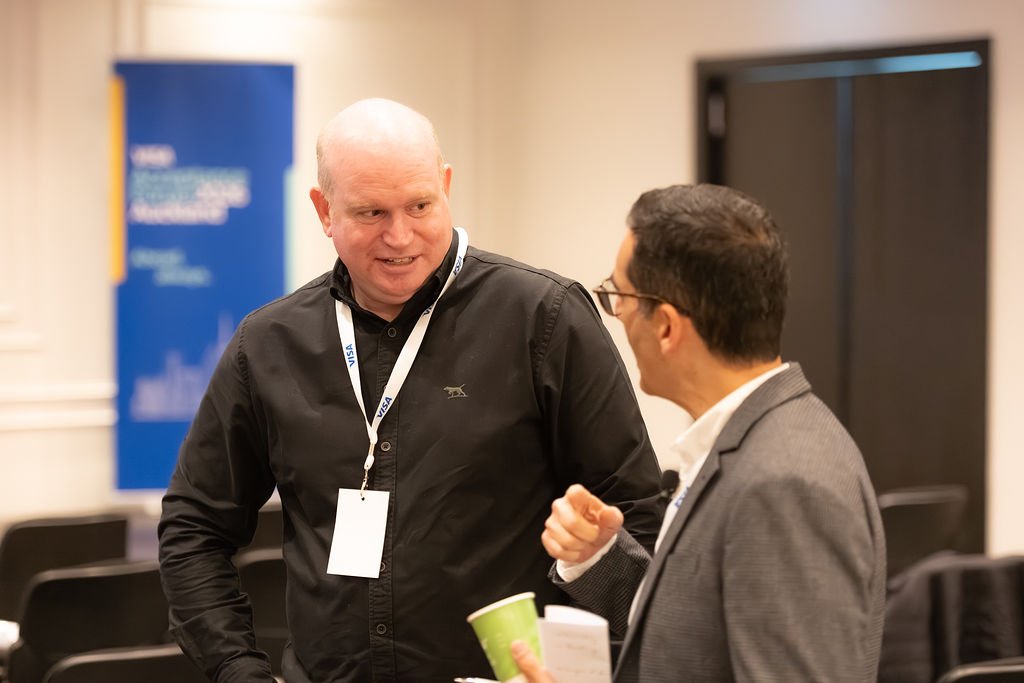 Two men engaged in conversation at a professional event, with conference badges around their necks. One man is bald and wearing a black shirt, while the other man has dark hair and glasses, dressed in a gray suit holding a paper cup and a notepad.