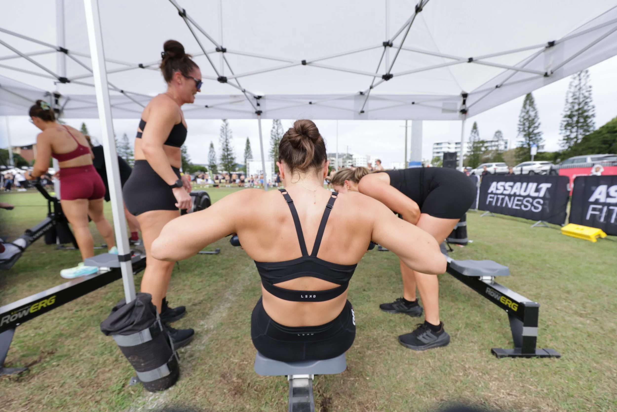 Women exercising on rowing machines under a white canopy tent at an outdoor fitness event with people and trees in the background.